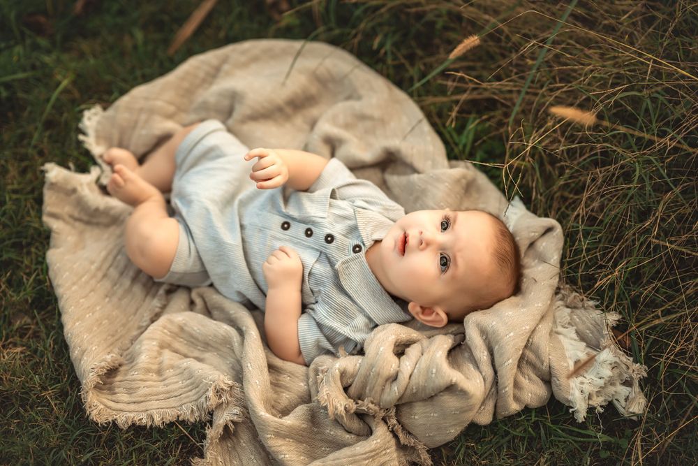 Baby wearing light blue outfit lies on a tan blanket in the grass while gazing upward for 6-month milestone photo session