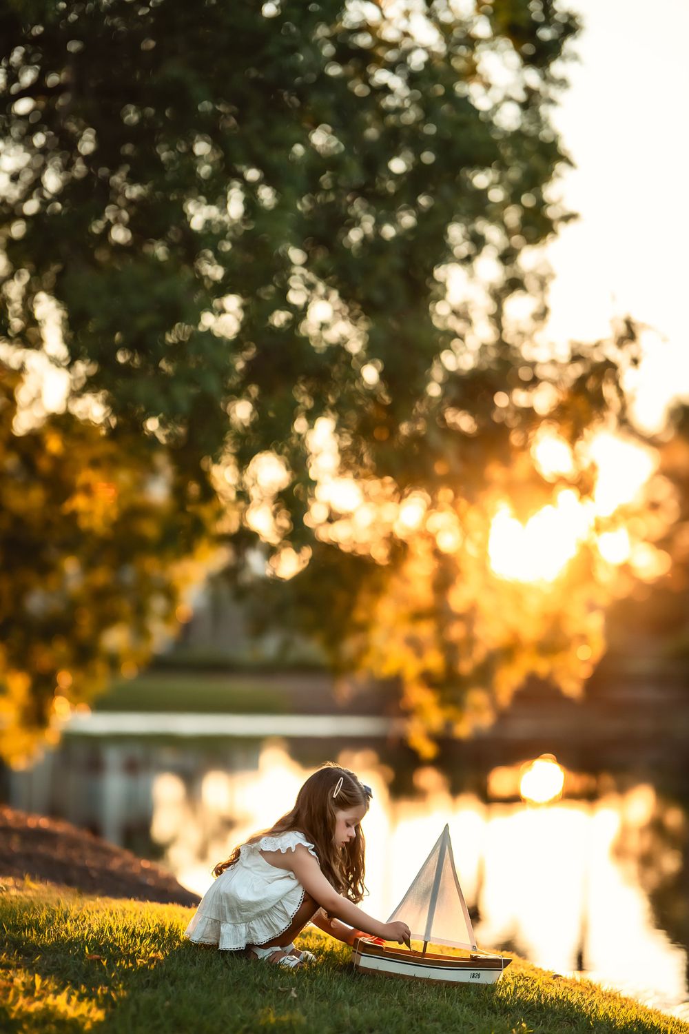 Child in white dress plays with toy boat by the lakeside during golden sunset creating magical summer evening atmosphere.