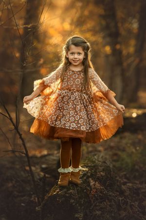 A child in a patterned daisy dress balances across a log against a golden autumn background.