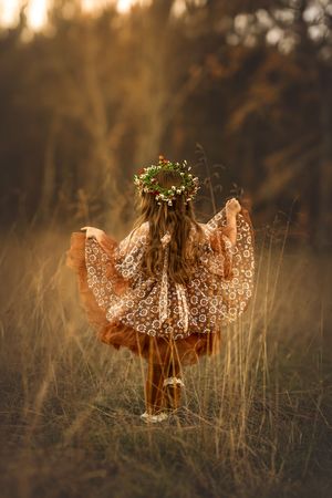 Young girl in orange daisy dress and flower crown frolicks in a field of tall golden grass.