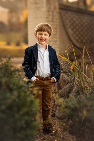 A child in navy blazer and brown pants stands confidently in an autumn garden.