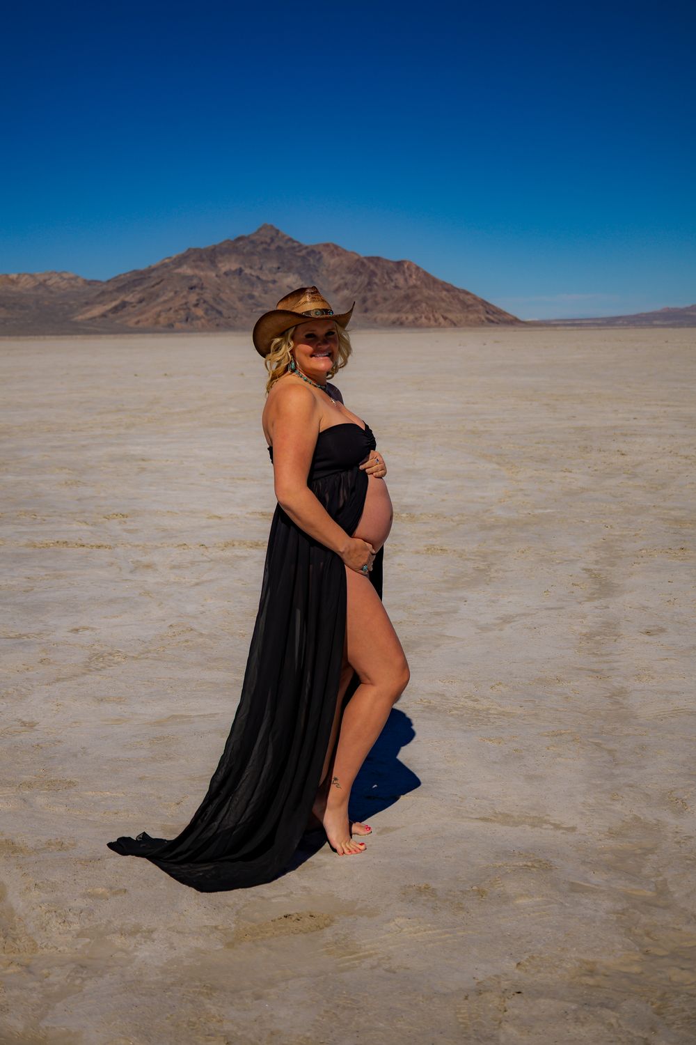 Pregnant woman in black gown and cowboy hat posing in salt flats desert with mountain backdrop.