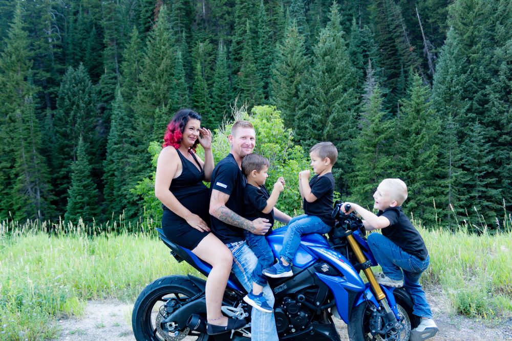 Group poses with a blue motorcycle against a backdrop of evergreen trees in the mountains.