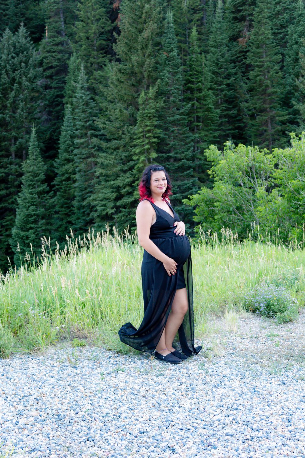 Woman in black dress poses on rocky ground with pine forest backdrop.