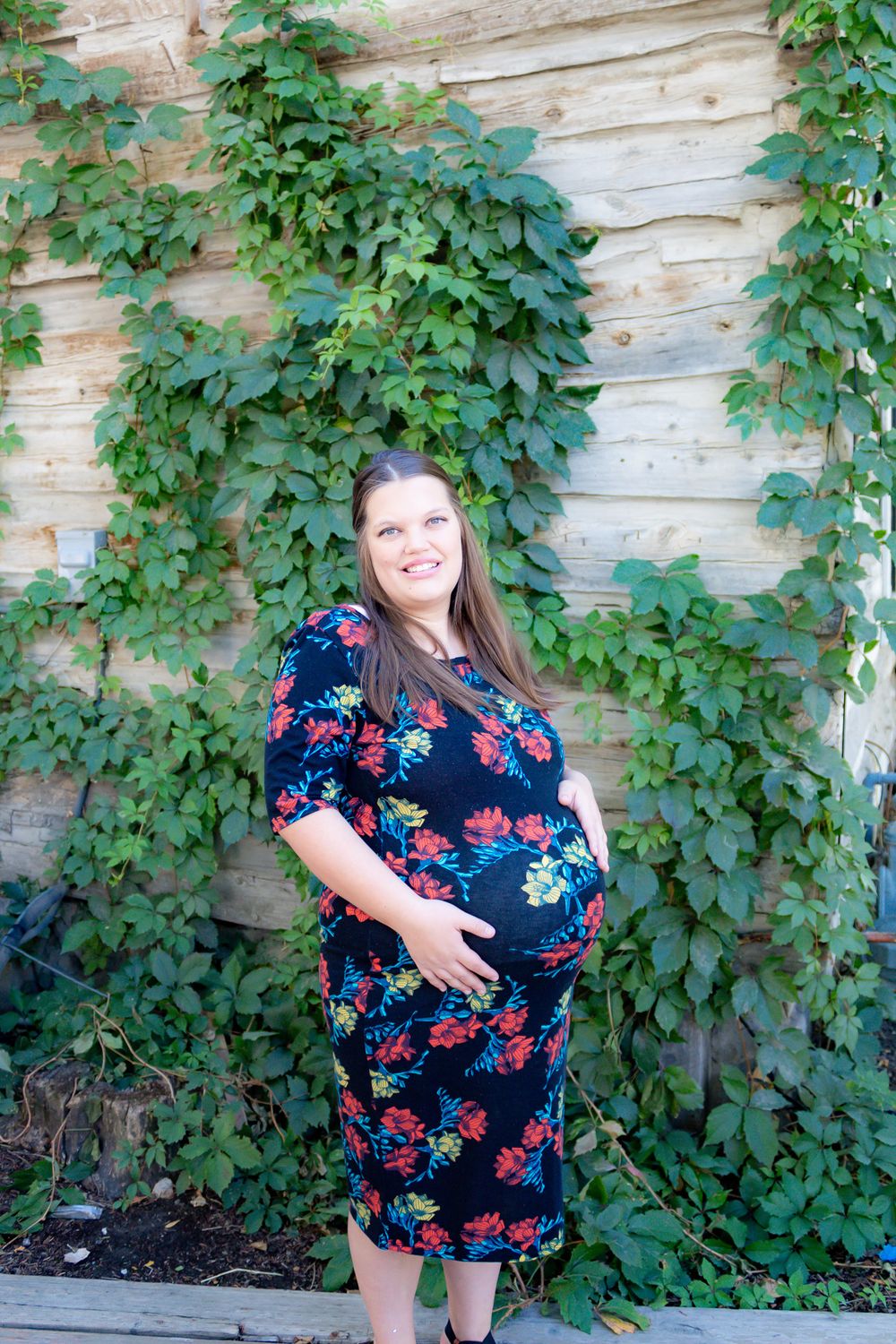 A woman in a floral dress poses against a wall covered in green ivy vines.