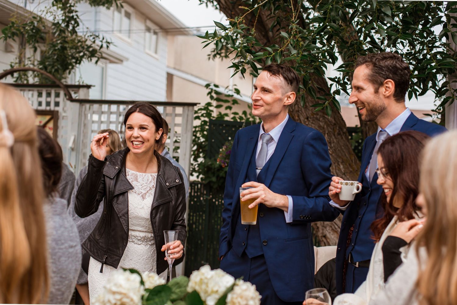 Casual outdoor gathering with laughing guests enjoying drinks under garden tree.