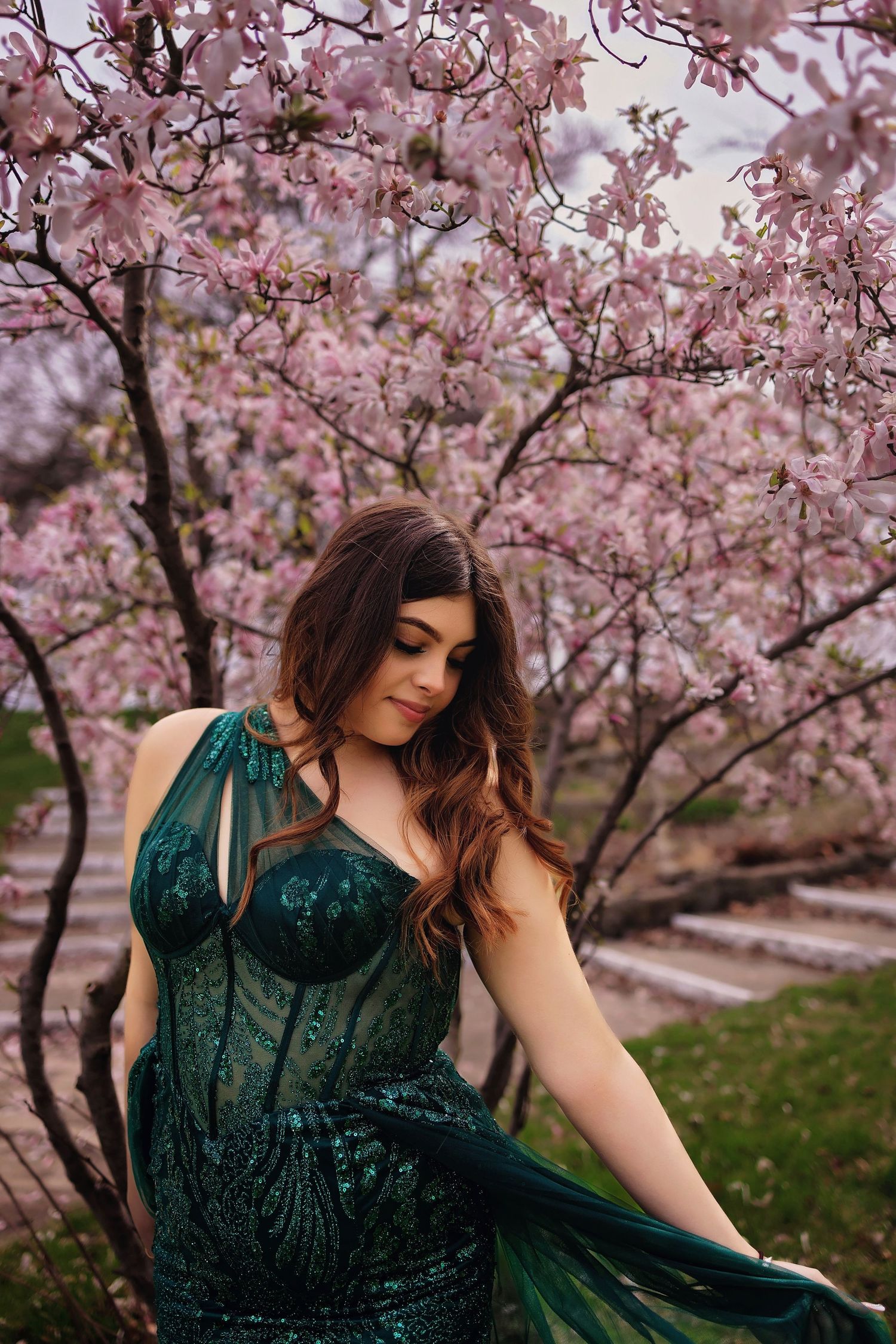 Woman in emerald green dress poses under blooming cherry blossom trees in spring.