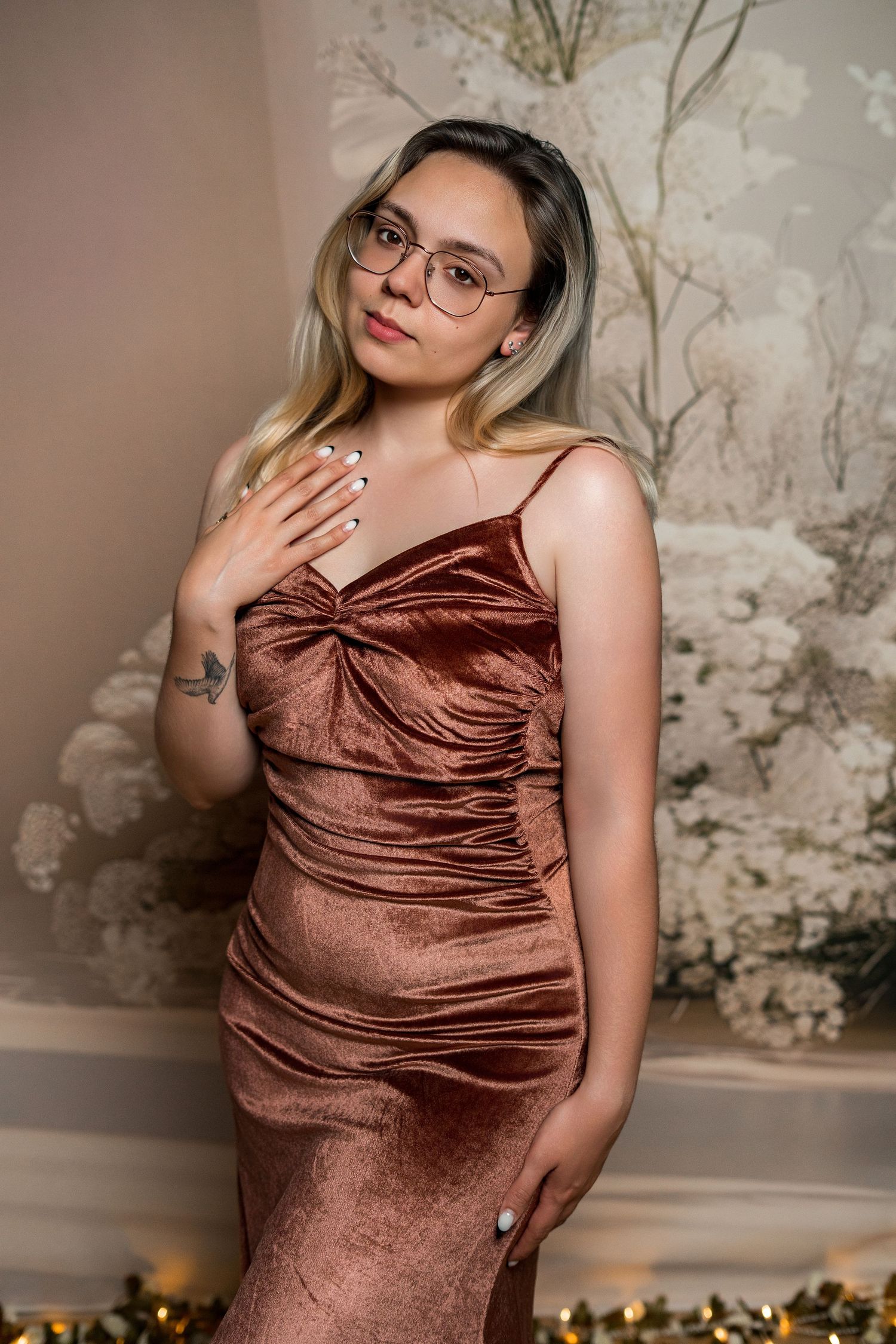 Woman in bronze velvet dress and glasses poses elegantly before floral backdrop with holiday lights.