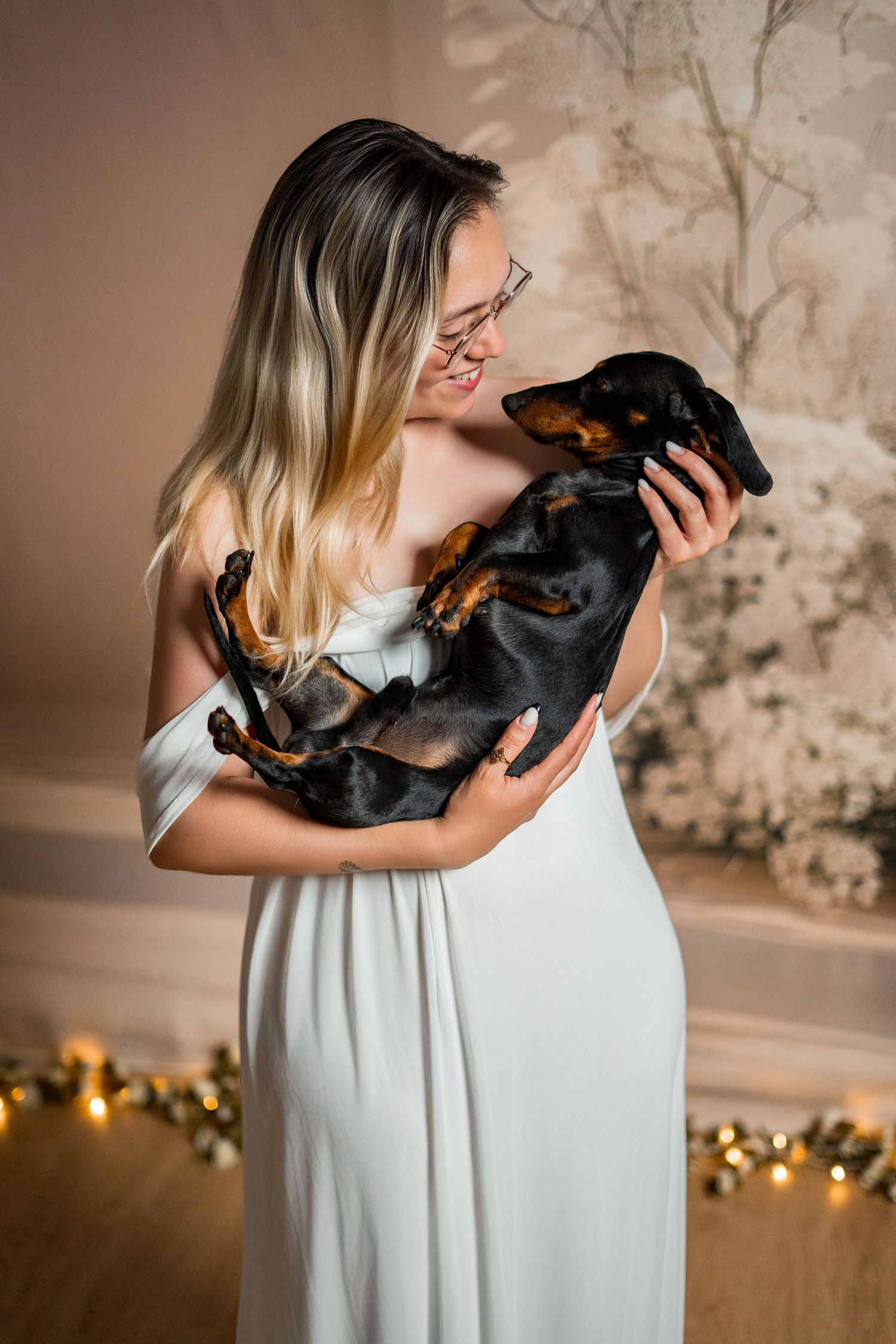 Smiling blonde woman in white dress holding a black and tan dachshund dog in a decorated room.