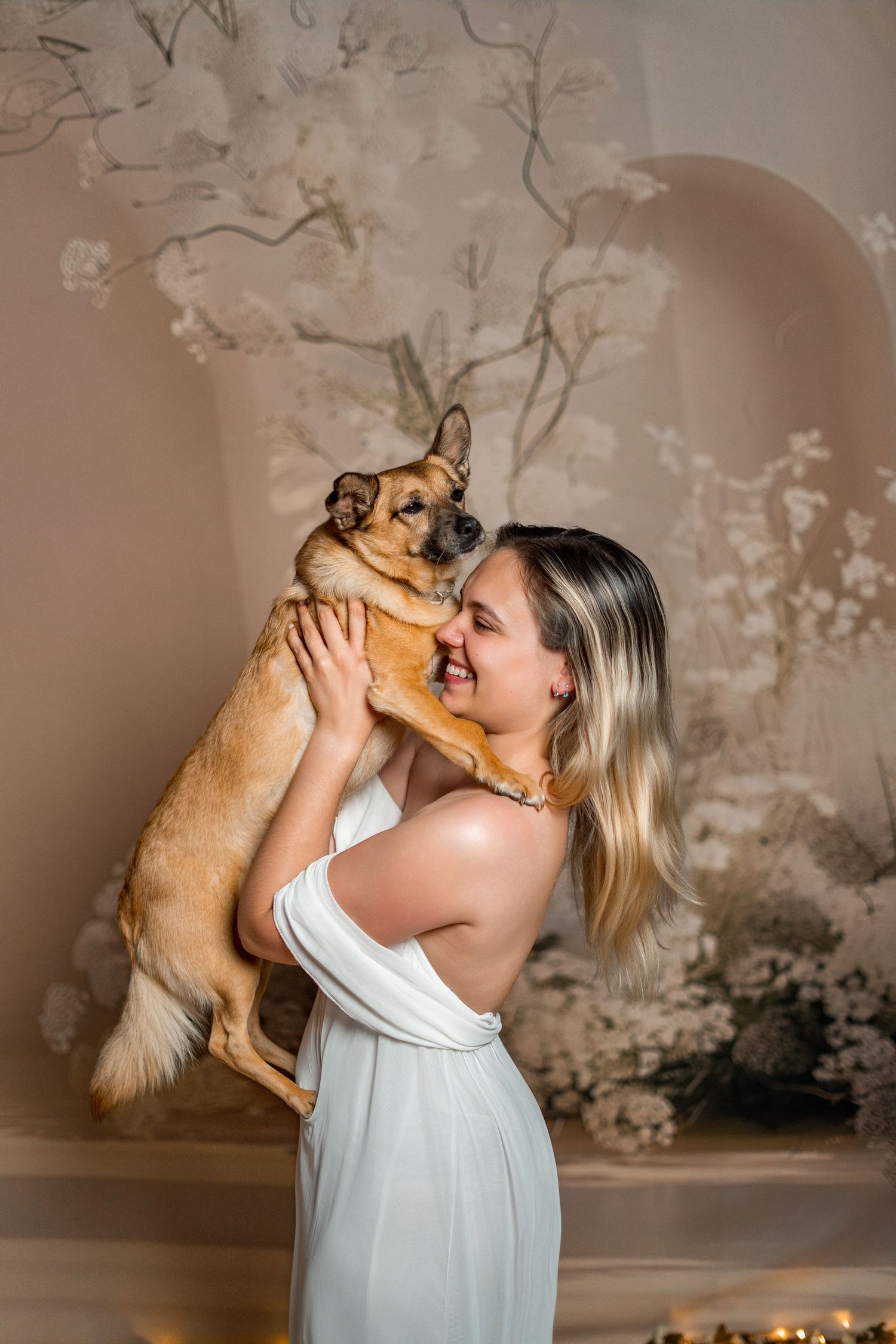 Woman in white dress joyfully holding a small brown dog up to her face in an elegant floral setting.