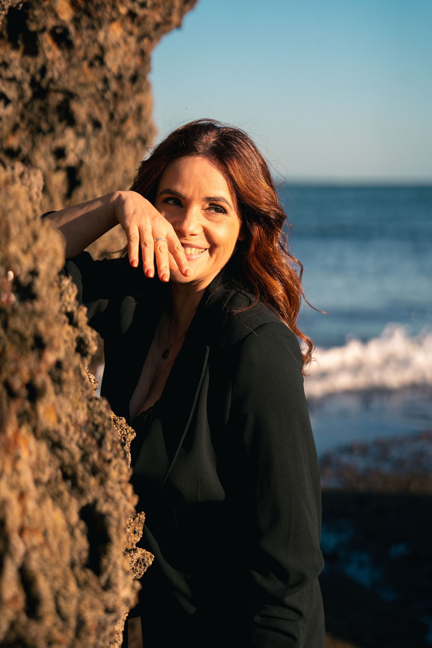 Woman in black sweater laughing against rocky beach backdrop at golden hour.