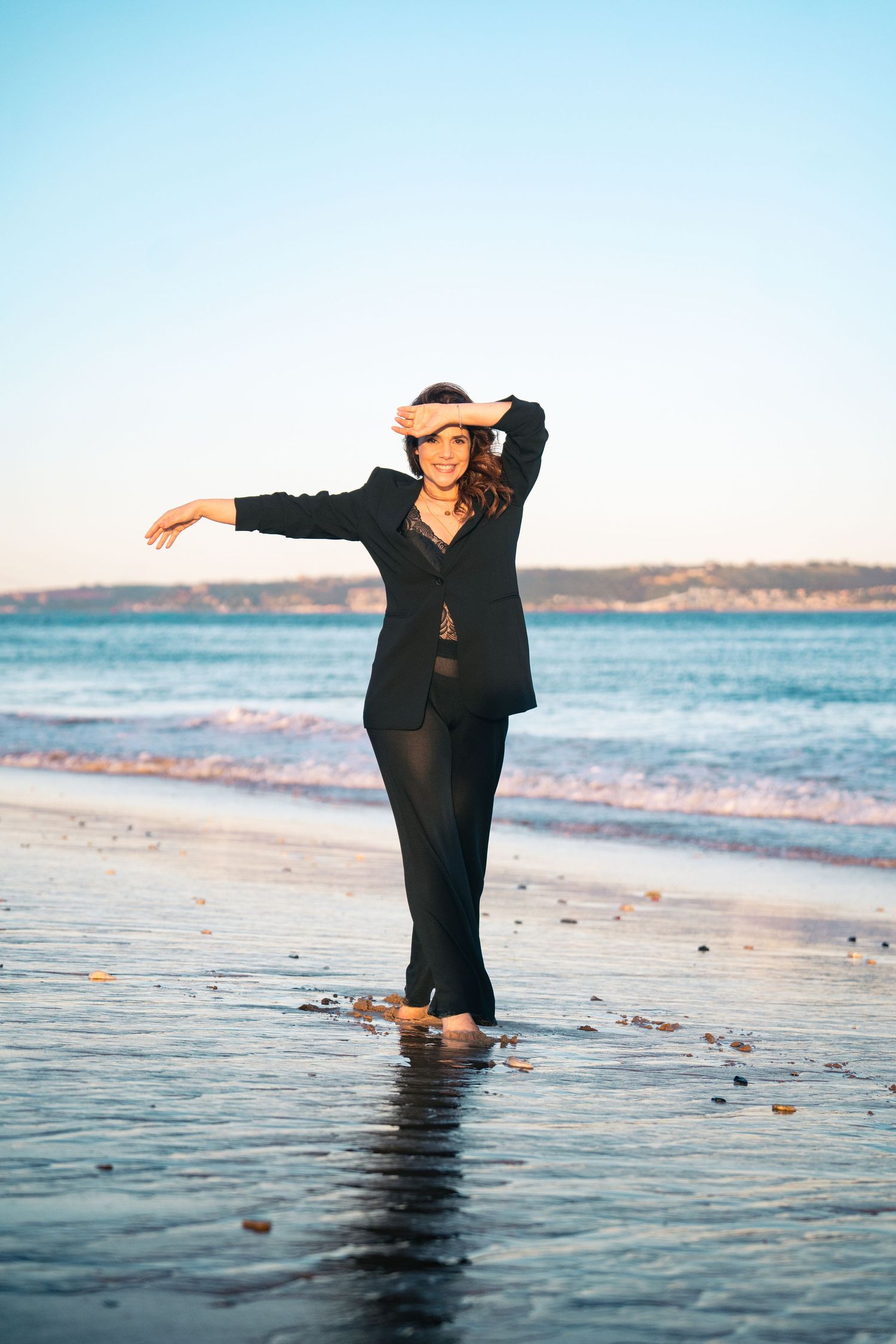 Woman in flowing black dress dancing in shallow ocean water at sunset.
