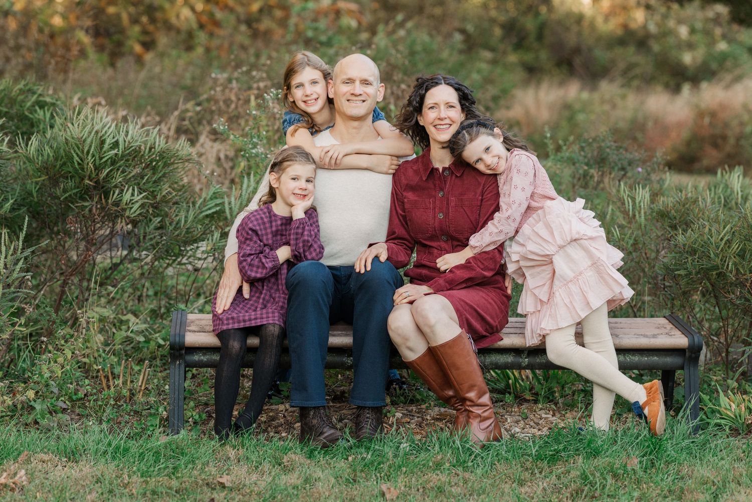 A family stands outdoors by a pond with their speckled gray dog in an autumn setting with colorful fall foliage.
