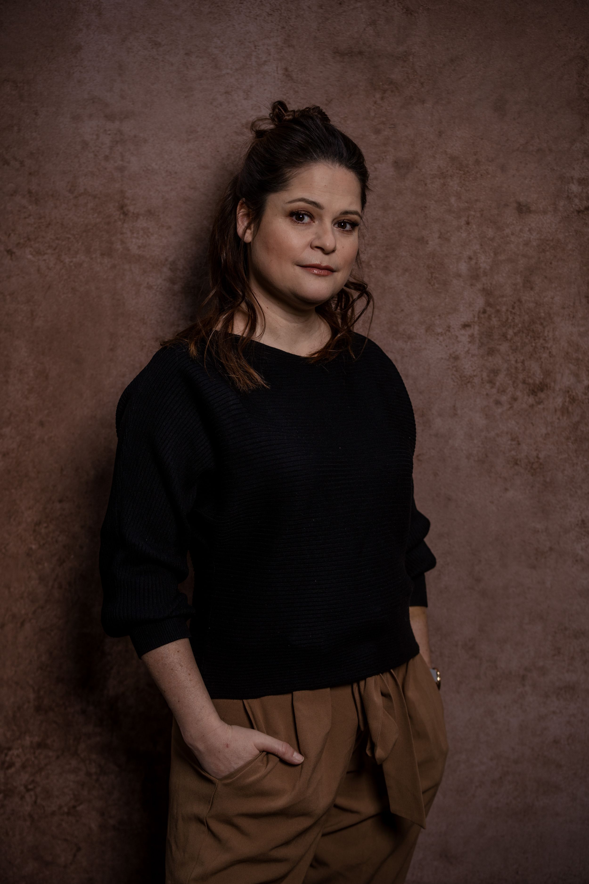 A portrait against a brown textured background showing someone in a black sweater and brown pants with hands in pockets.