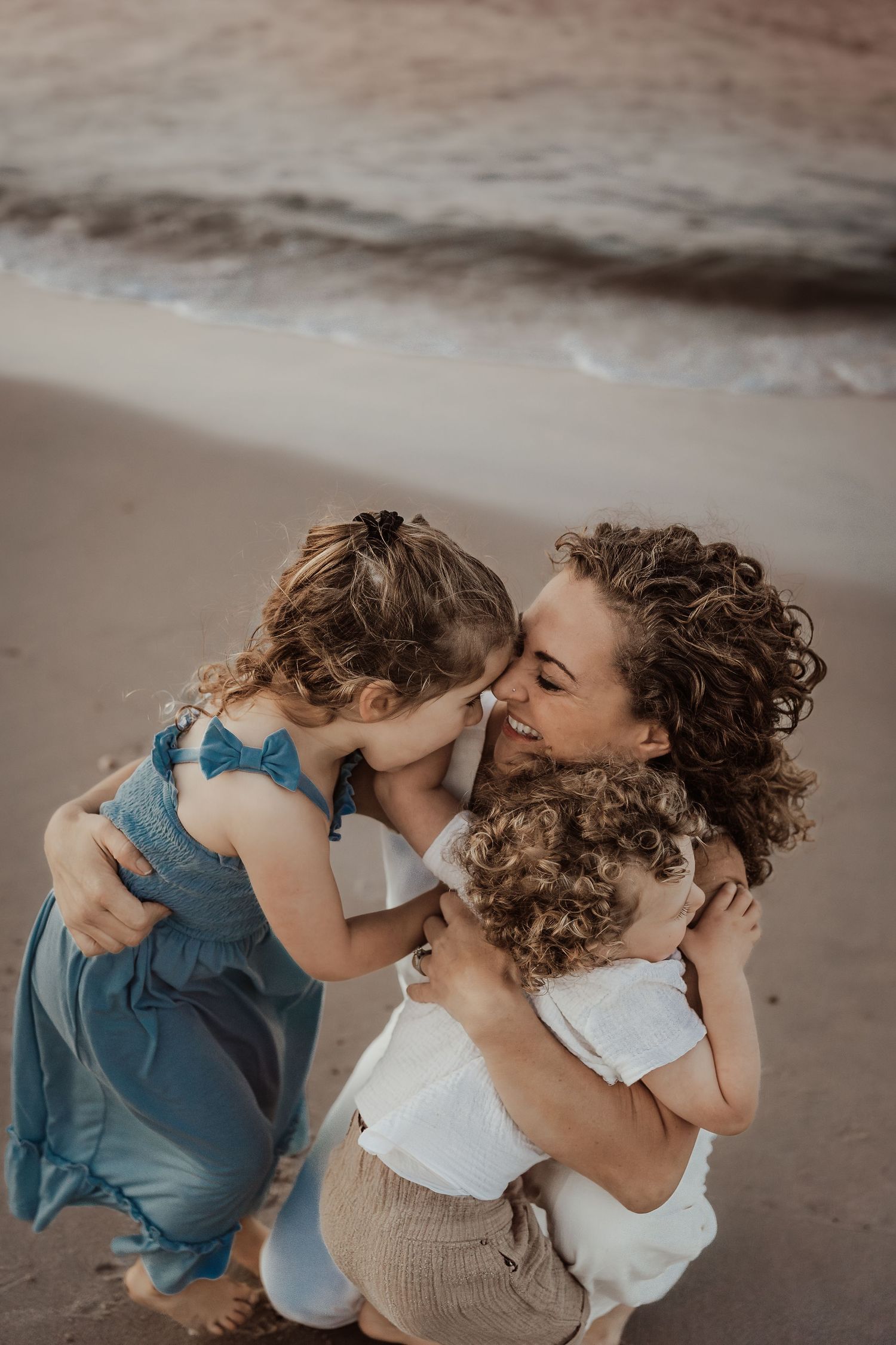 Mother with curly hair and her two young children embracing on a Delaware beach being photographed from above