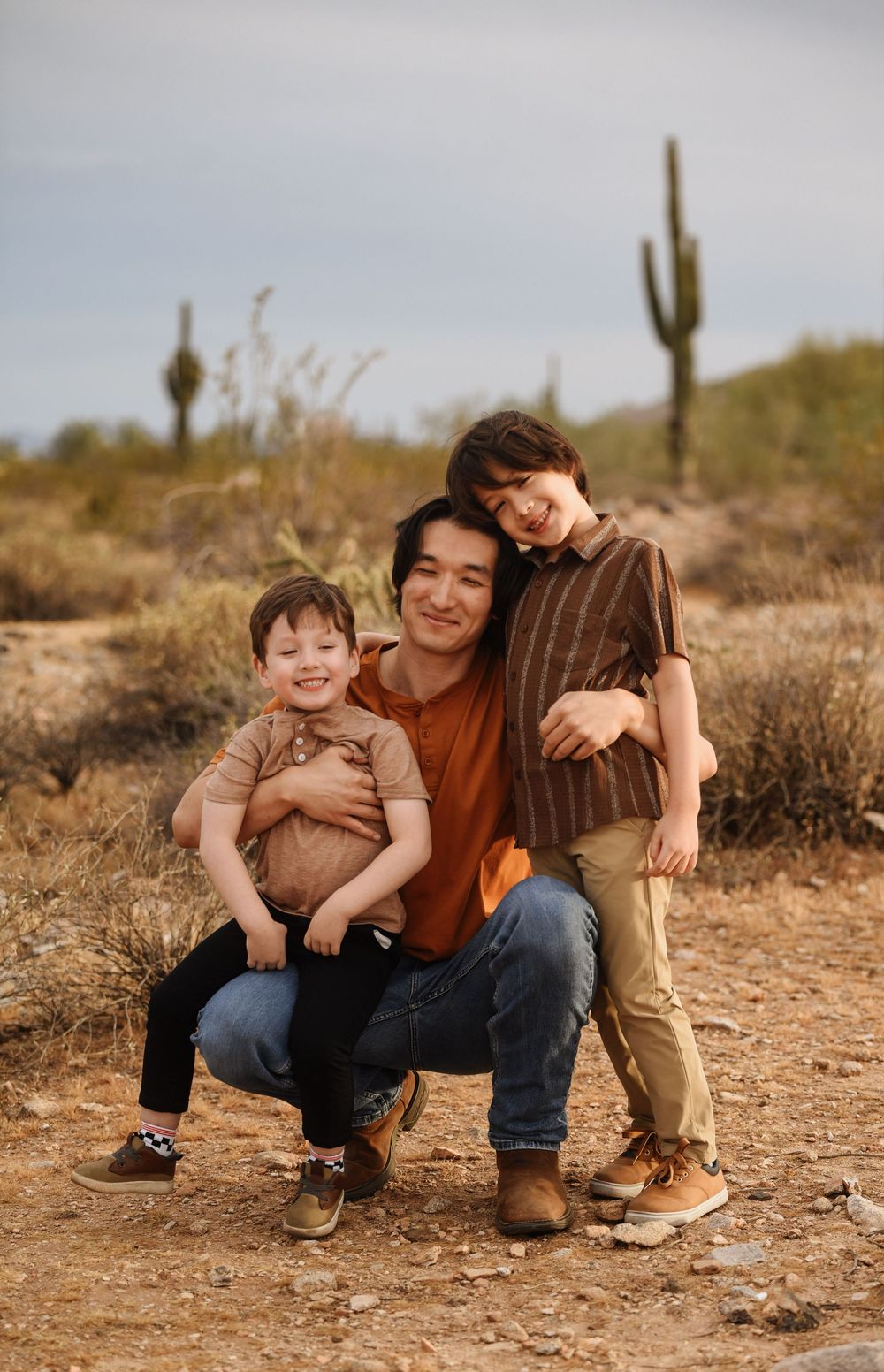 A father crouches in a desert landscape with two young sons, surrounded by saguaro cacti under a cloudy sky.