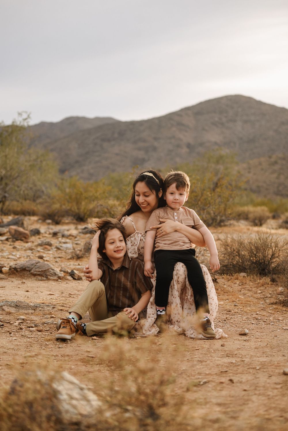 A mother sits on desert ground with two young children, mountains and arid landscape in background.