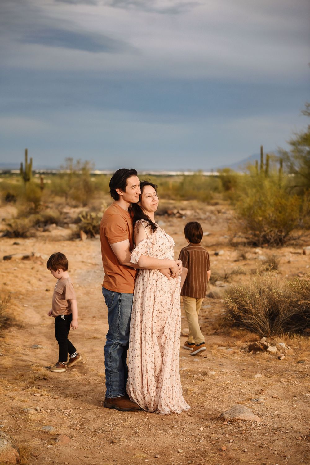 A couple embraces in an Arizona desert landscape with saguaro cacti while two young children play nearby.