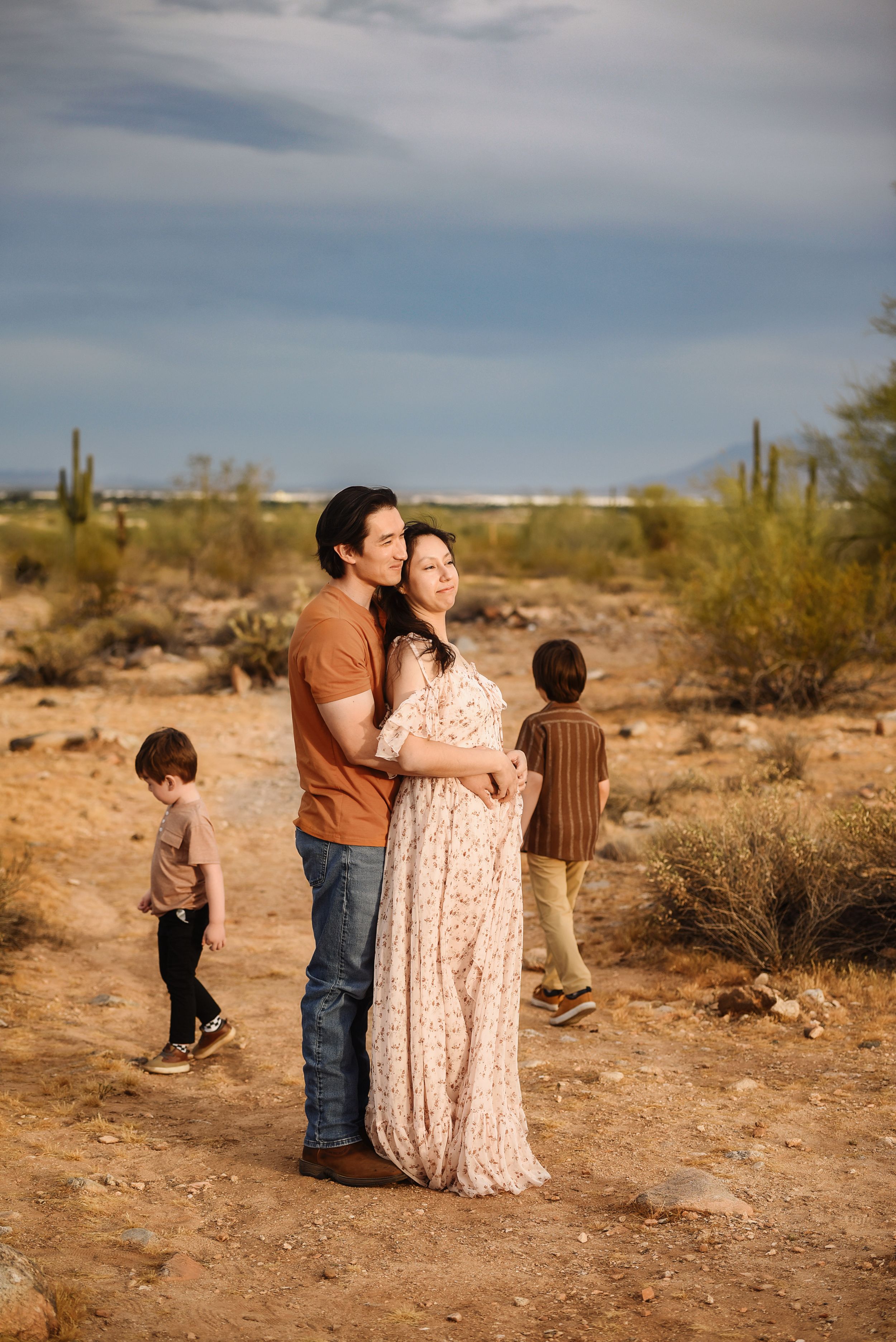 A couple embraces in an Arizona desert landscape with saguaro cacti while two young children play nearby.