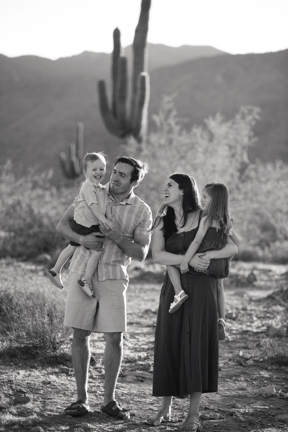 Black and white family photo in Arizona desert with saguaro cactus, parents holding two young children, smiling outdoors.