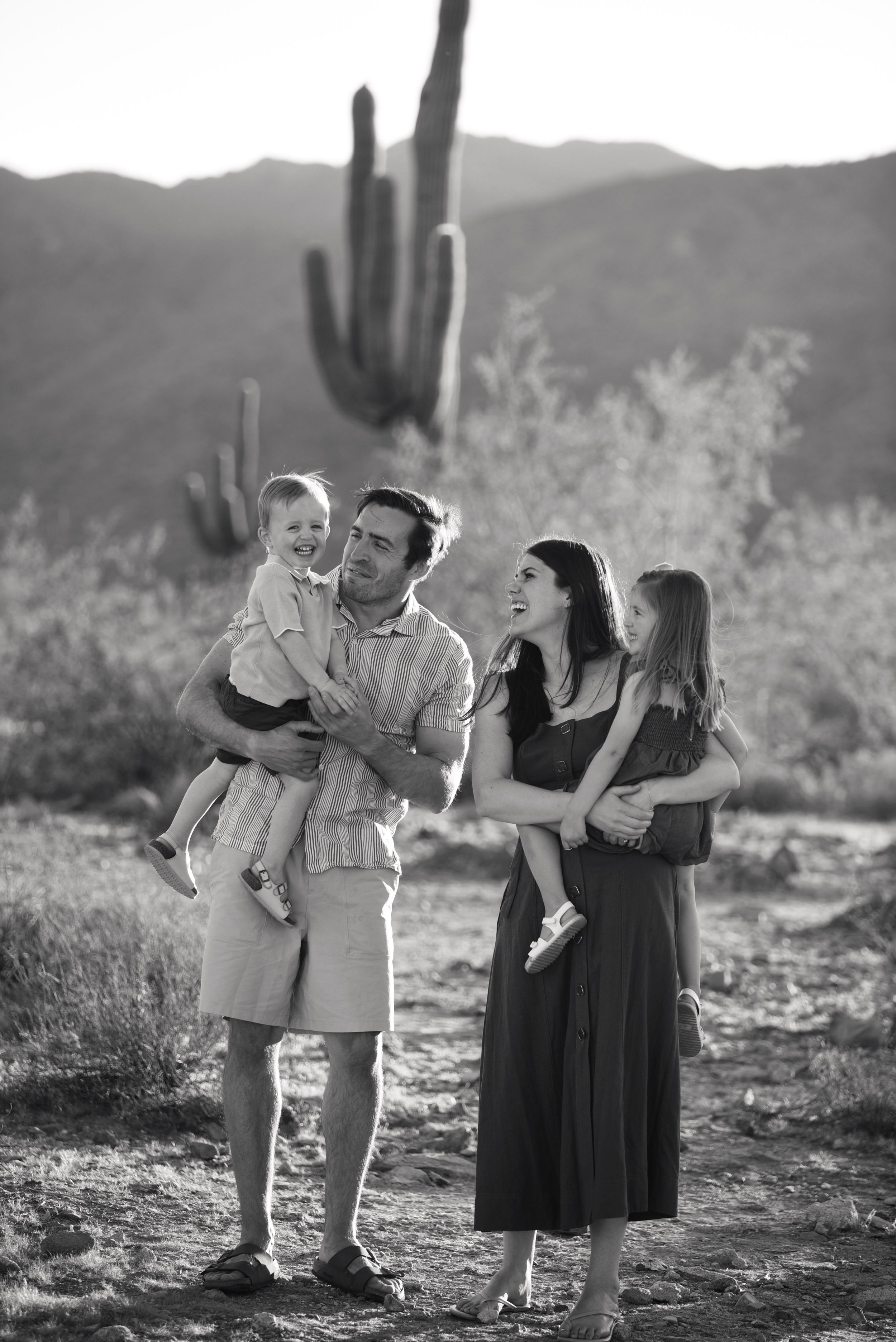 Black and white family photo in Arizona desert with saguaro cactus, parents holding two young children, smiling outdoors.