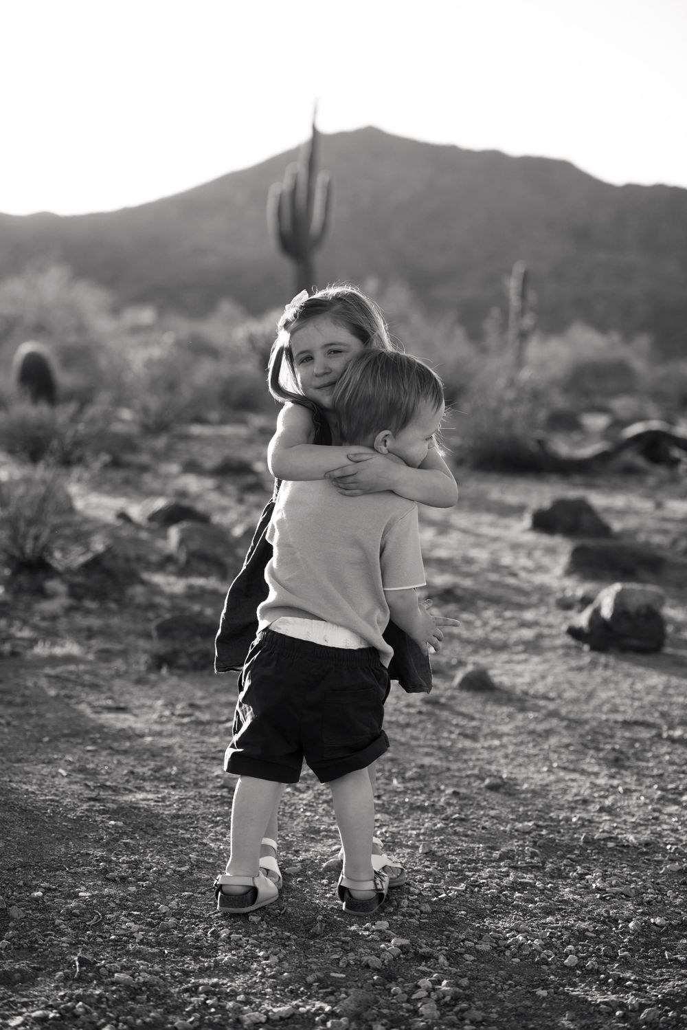 Two children hugging in a desert landscape with saguaro cacti and mountains in black and white photo.