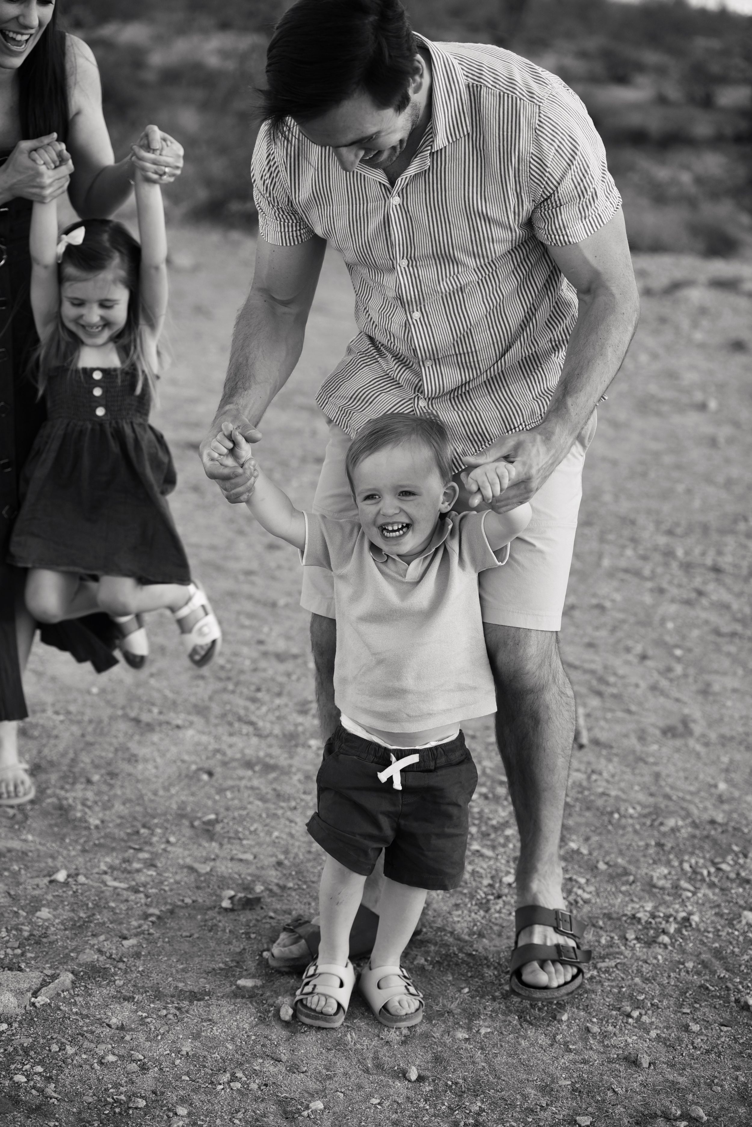 Black and white photo of a laughing toddler boy being helped to walk by his father during a family outdoor photo session.