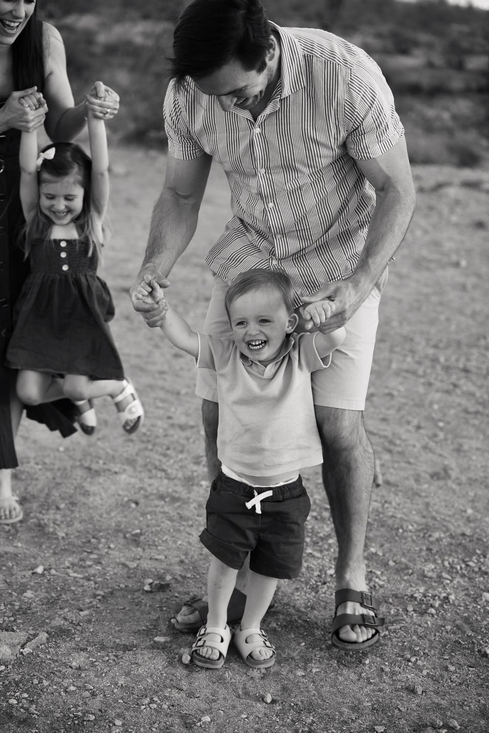 Black and white photo of a laughing toddler boy being helped to walk by his father during a family outdoor photo session.