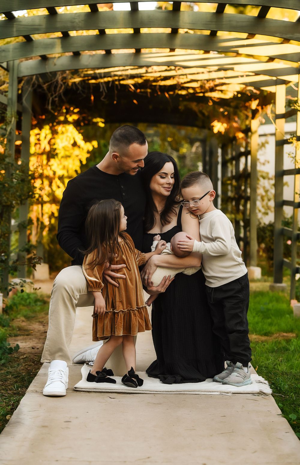 Family of four posing under a pergola with warm golden lighting, parents holding infant while young children gather close.
