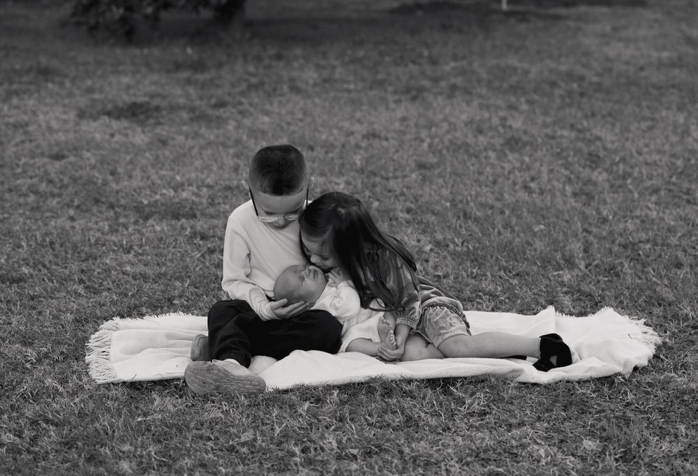 Black and white photo of two children sitting on a blanket outdoors, lovingly holding a newborn baby.