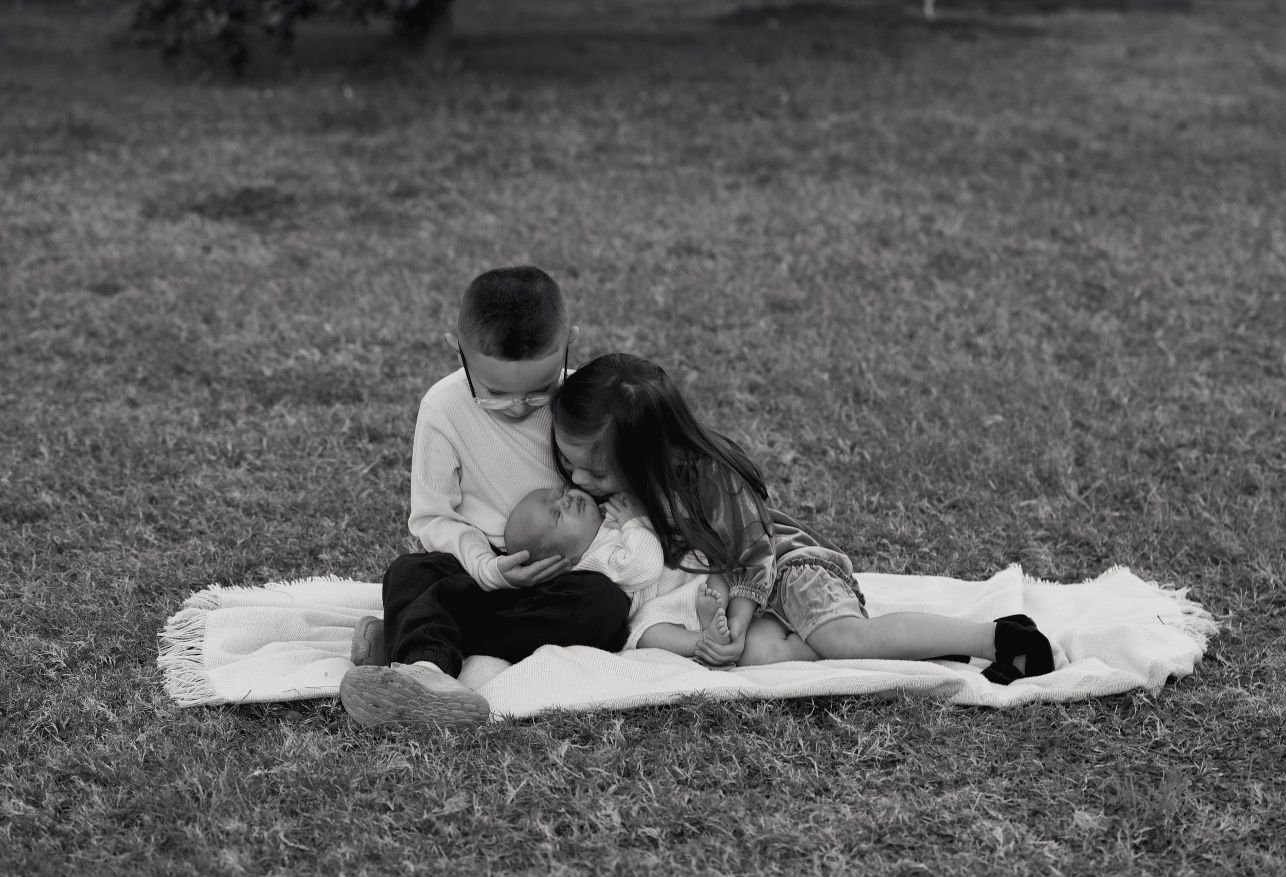 Black and white photo of two children sitting on a blanket outdoors, lovingly holding a newborn baby.