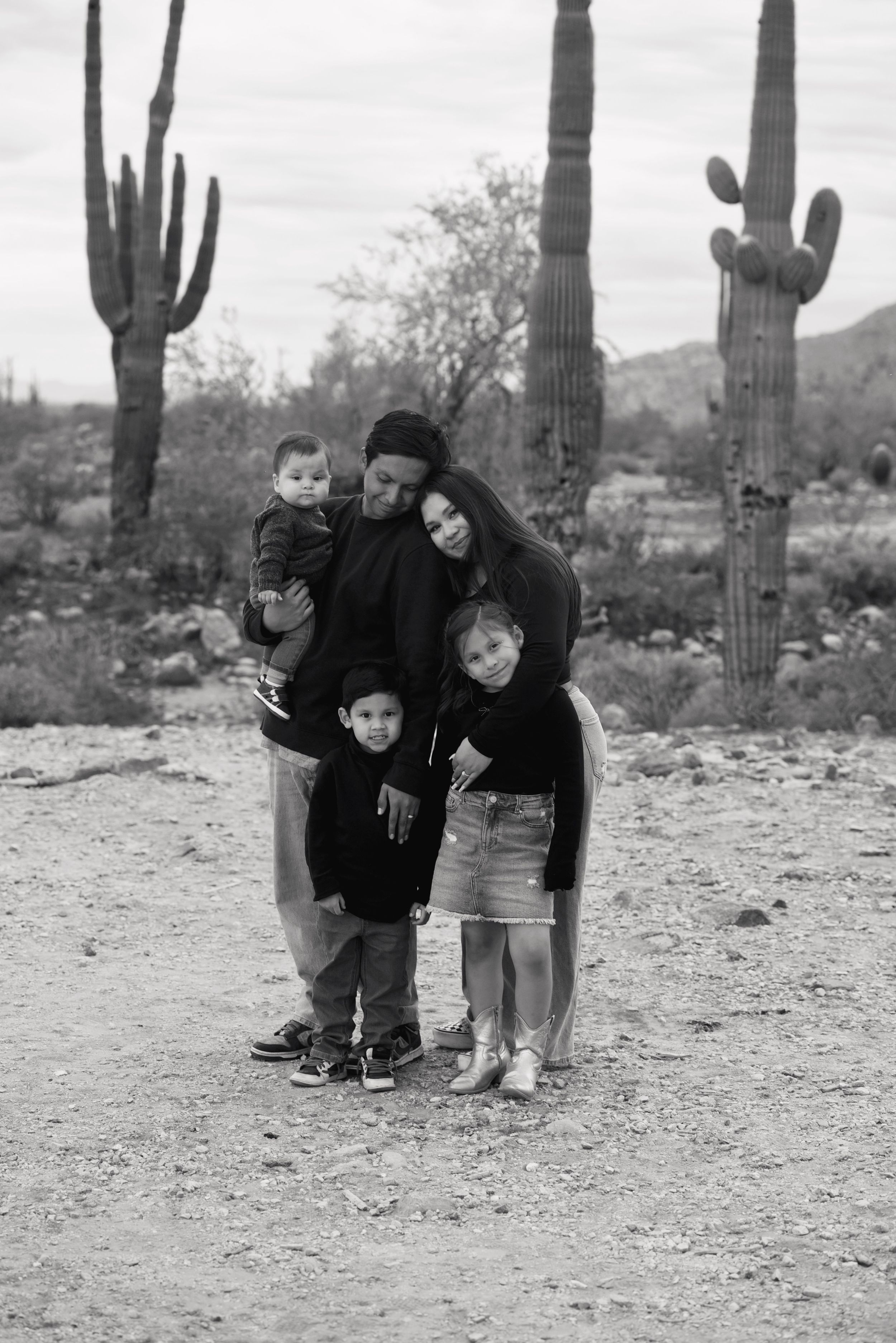 Black and white family portrait with saguaro cacti in Arizona desert, parents holding three young children.