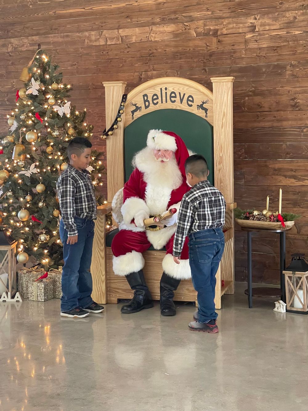 A child in plaid and jeans approaches Santa Claus seated in a rustic wooden room decorated with a Christmas tree.
