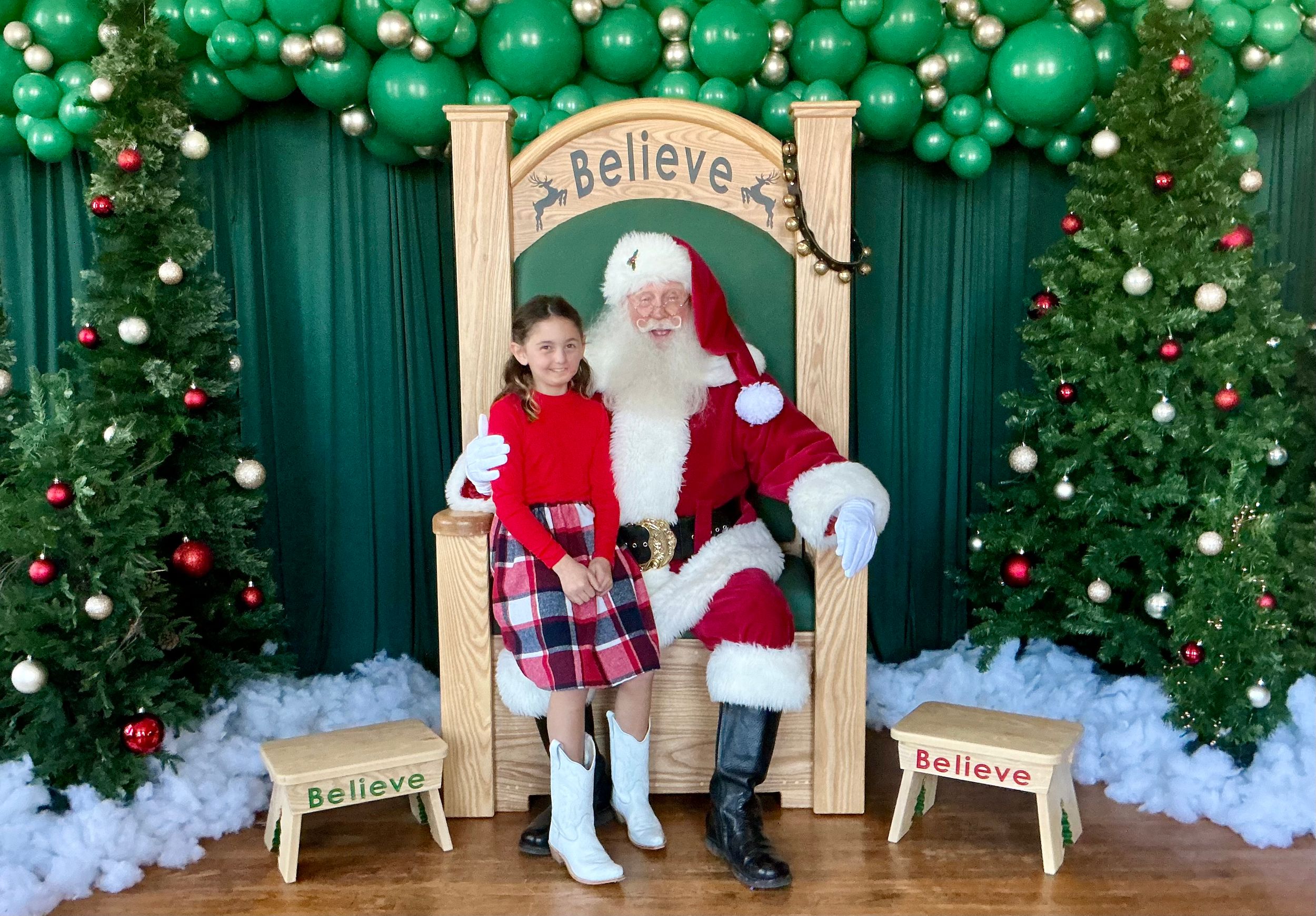 A young girl in a red dress poses with Santa Claus on a festive chair surrounded by Christmas trees and balloon arch.