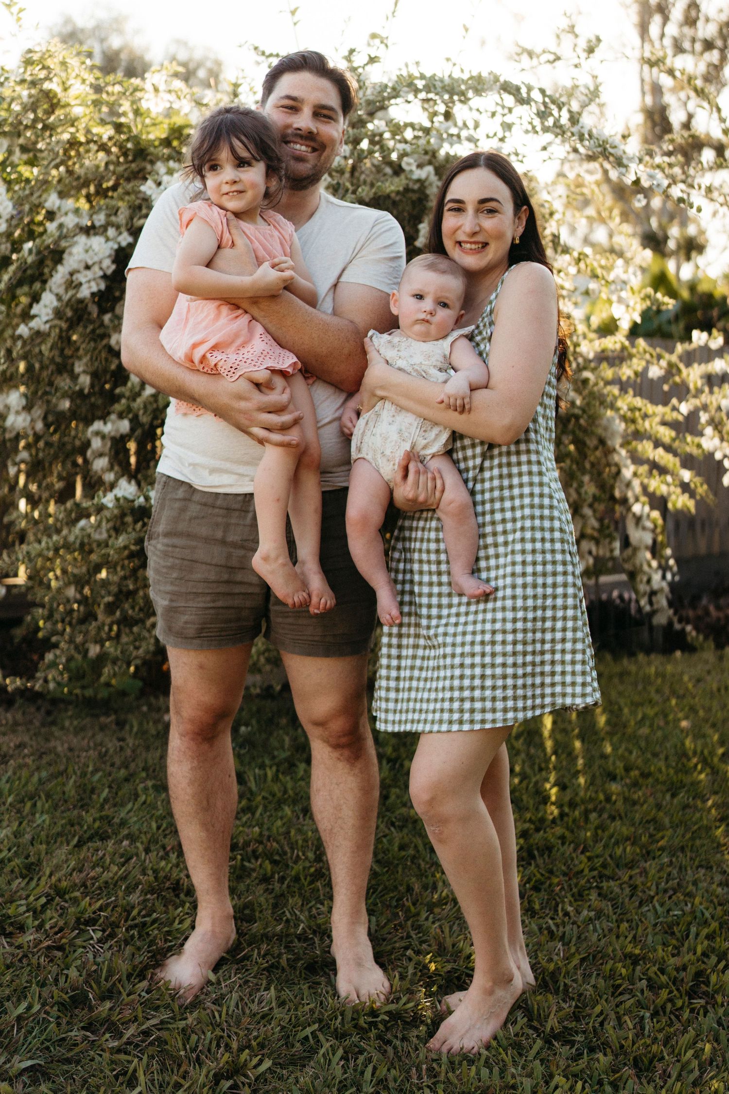 A happy family stands barefoot on grass during golden hour, with flowering trees visible in the background.