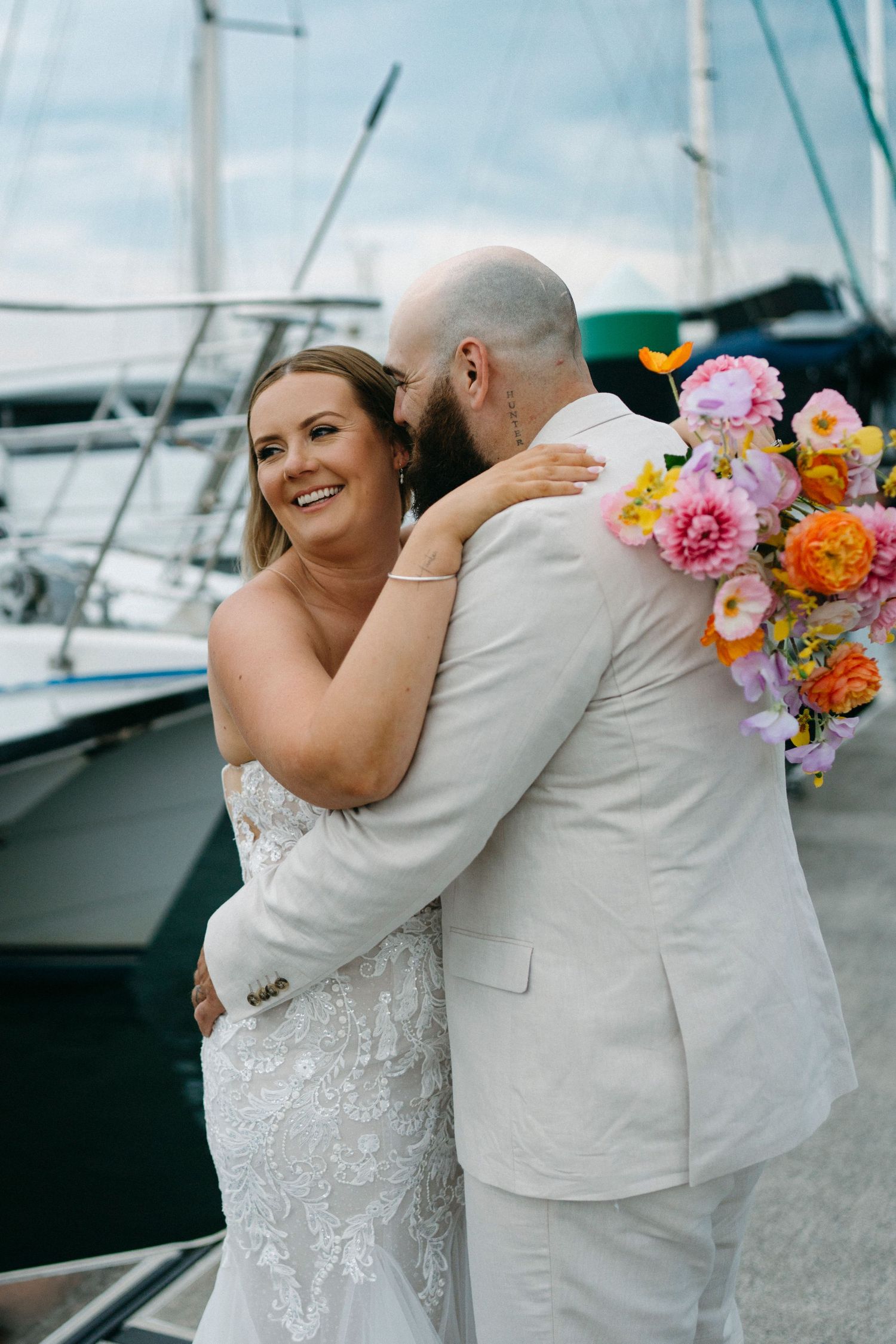 A couple embraces at a marina on their wedding day with colorful bouquet against backdrop of boats and blue sky.