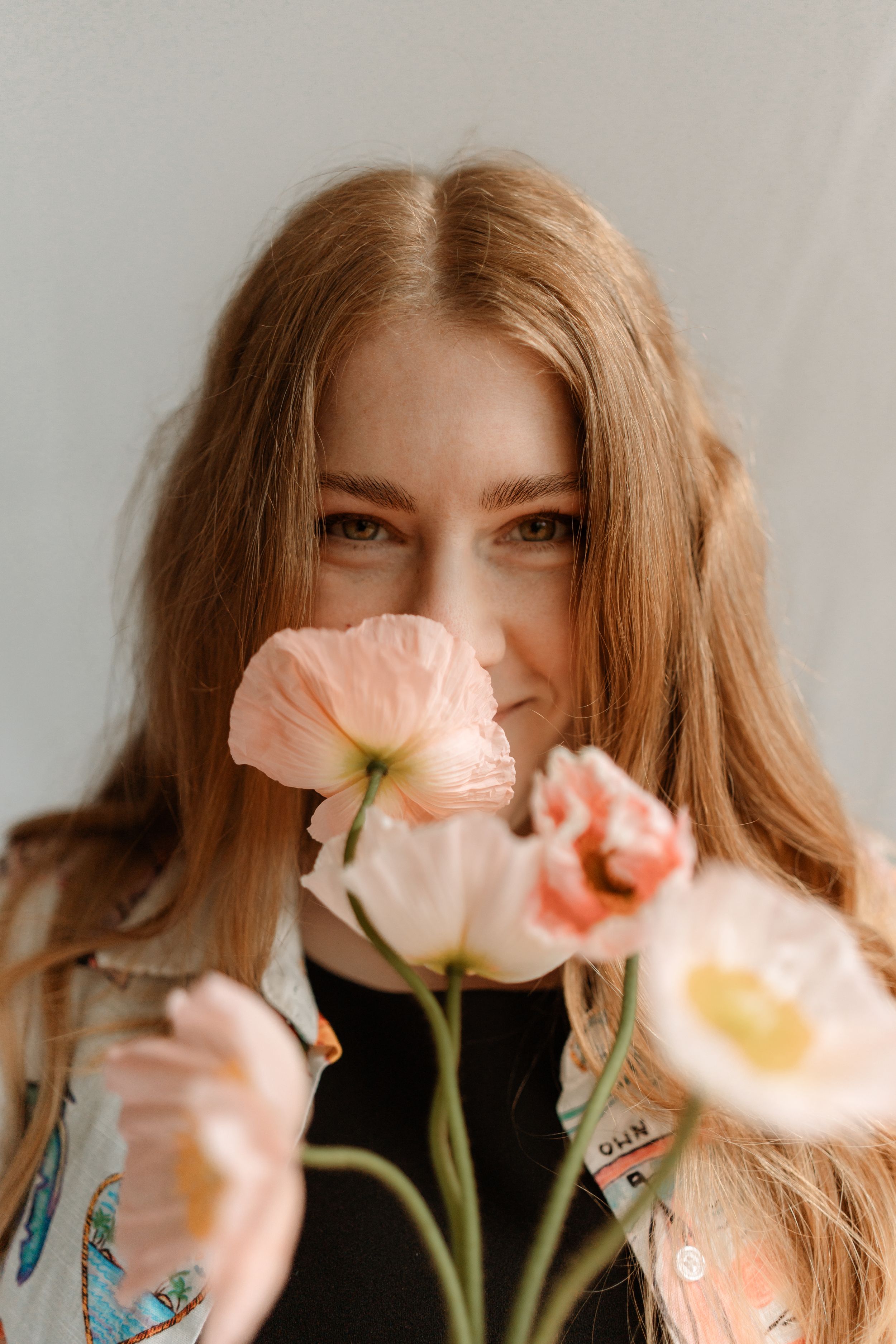 Young woman with long auburn hair holding soft pink poppy flowers close to her face against a light gray background.