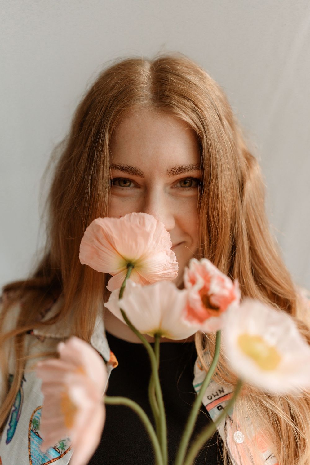 Young woman with long auburn hair holding soft pink poppy flowers close to her face against a light gray background.