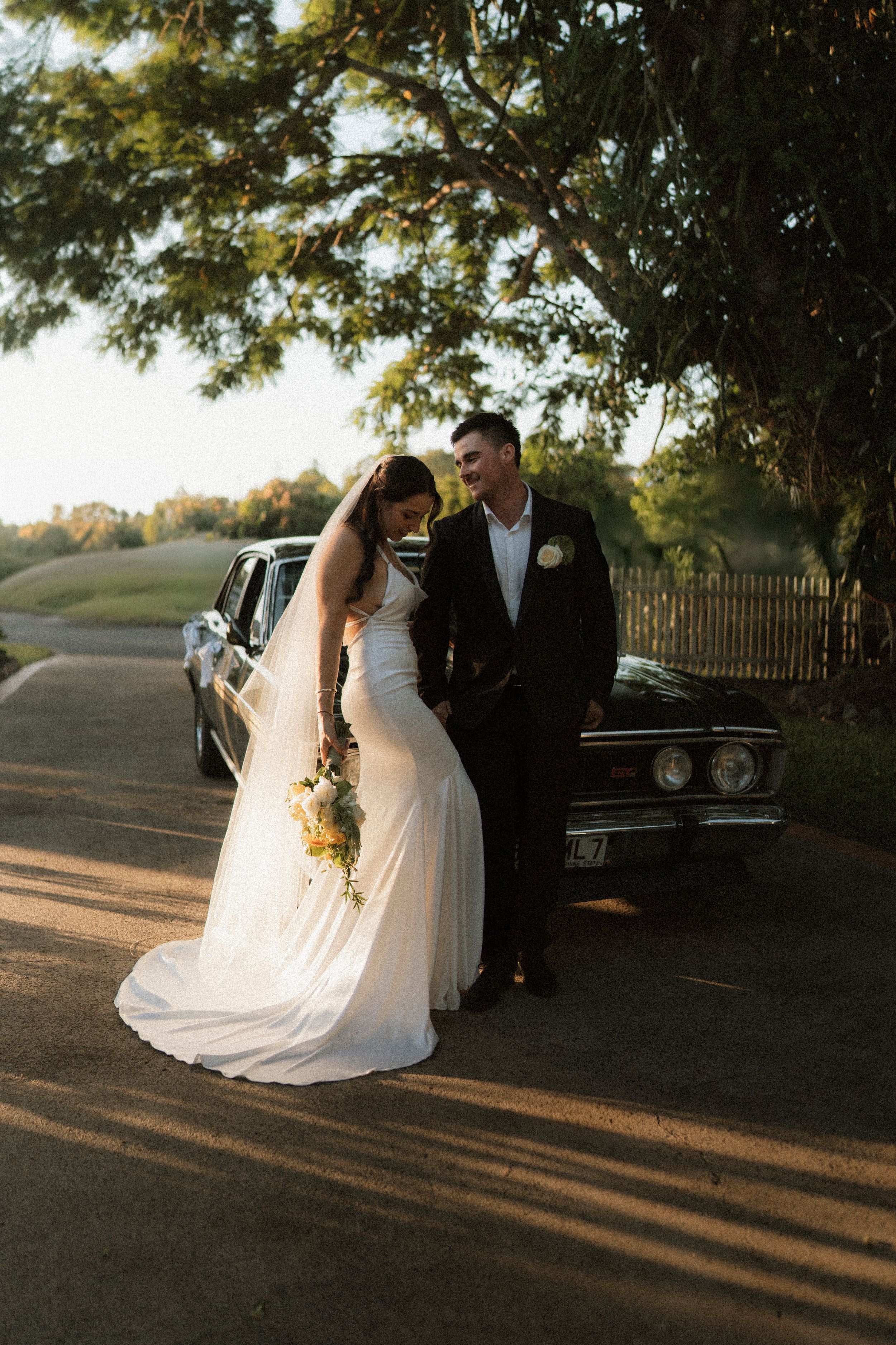 Bride and groom pose together in golden hour light beside a classic black car on a tree-lined driveway.
