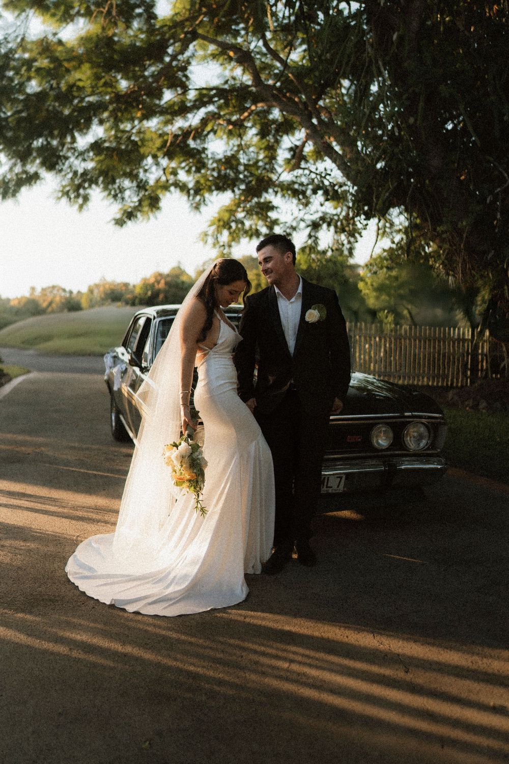 Bride and groom pose together in golden hour light beside a classic black car on a tree-lined driveway.