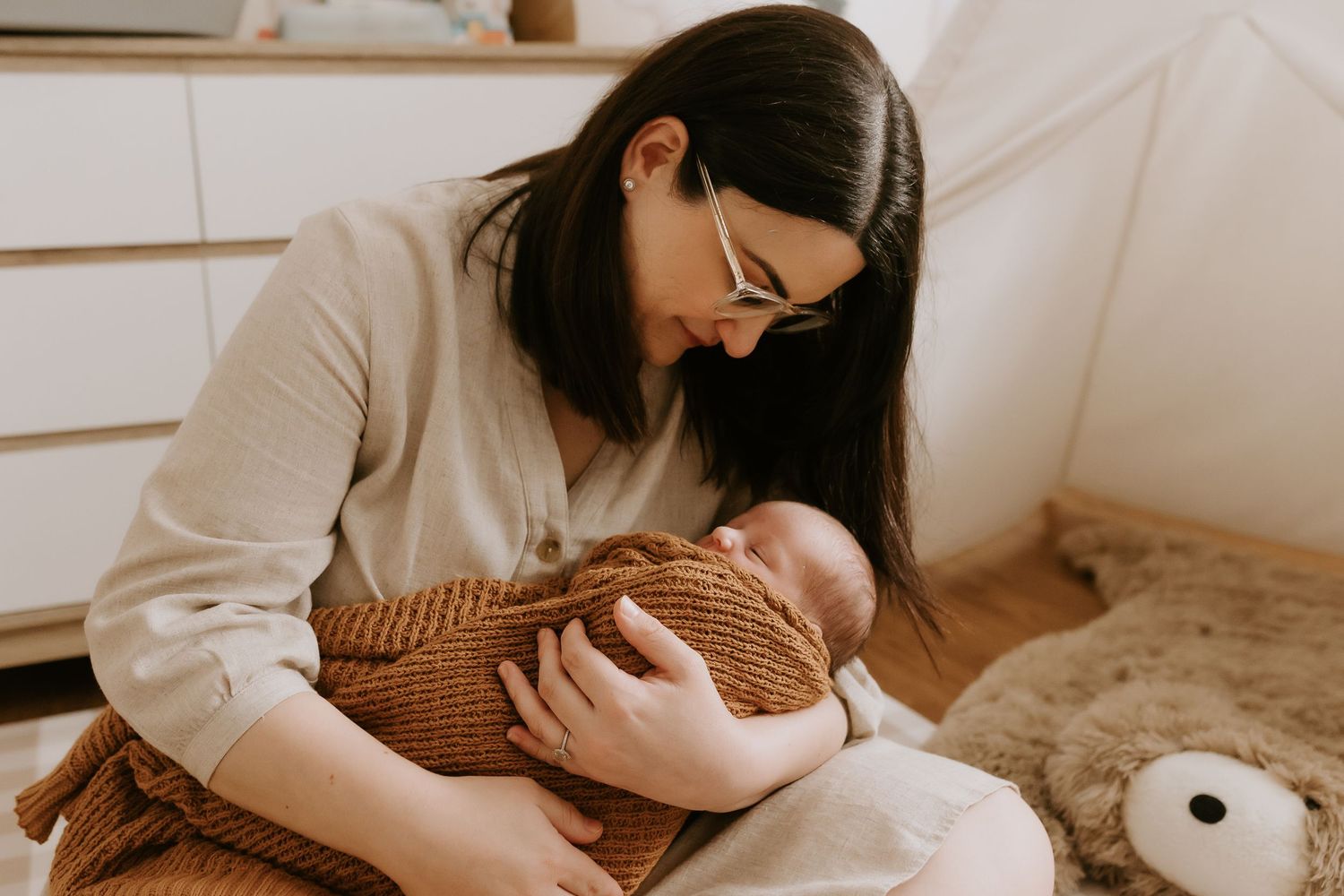 Woman in cream sweater tenderly holds infant wrapped in brown knit blanket in bright minimalist setting.