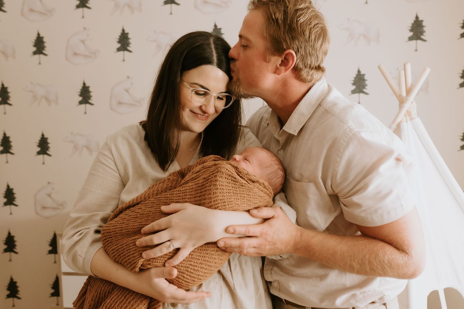 Couple shares intimate moment with newborn wrapped in brown blanket against tree-patterned wallpaper.
