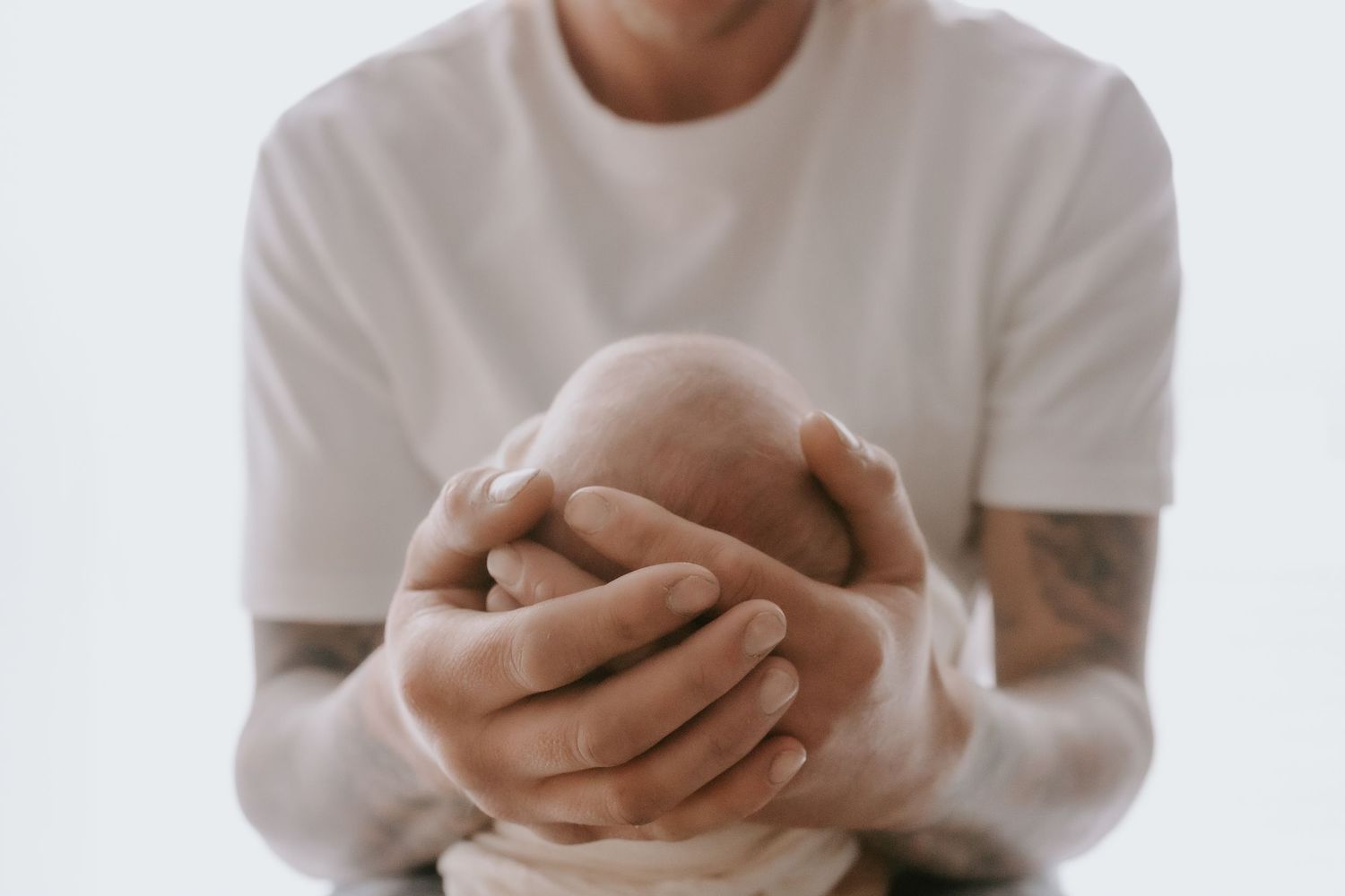 Tattooed hands gently cradle newborn baby against white background.