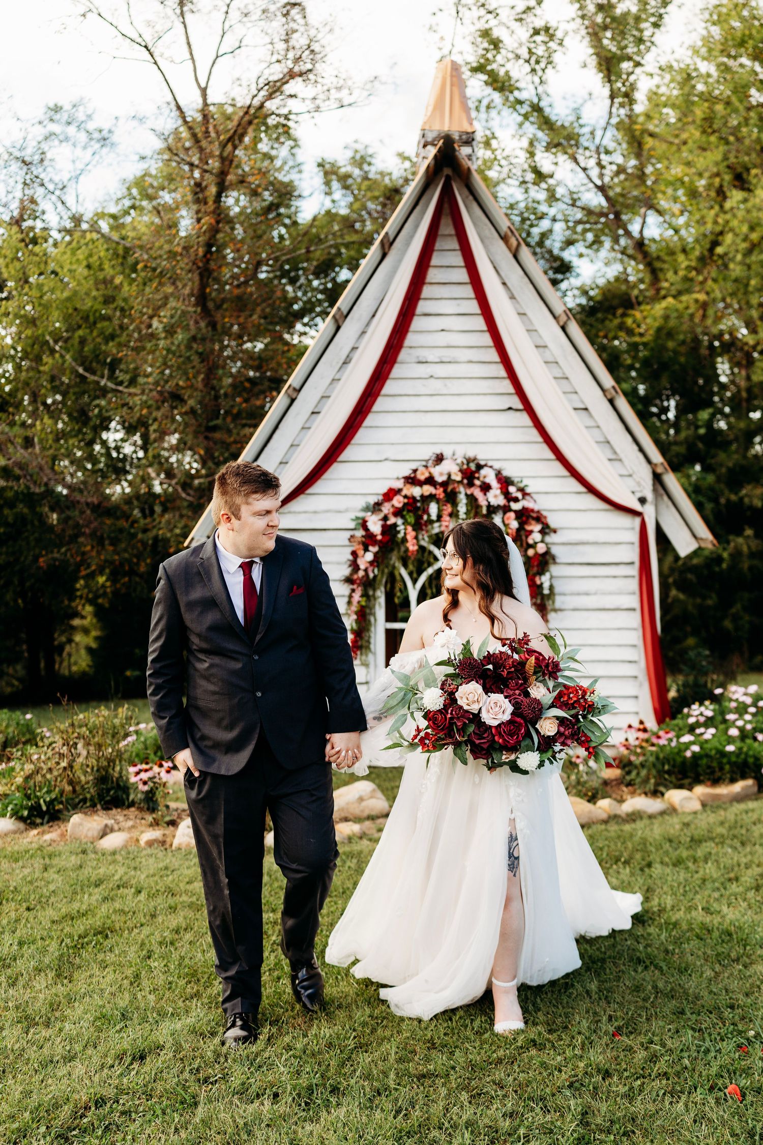 Bride and groom walk hand in hand before charming white chapel adorned with red floral arch.
