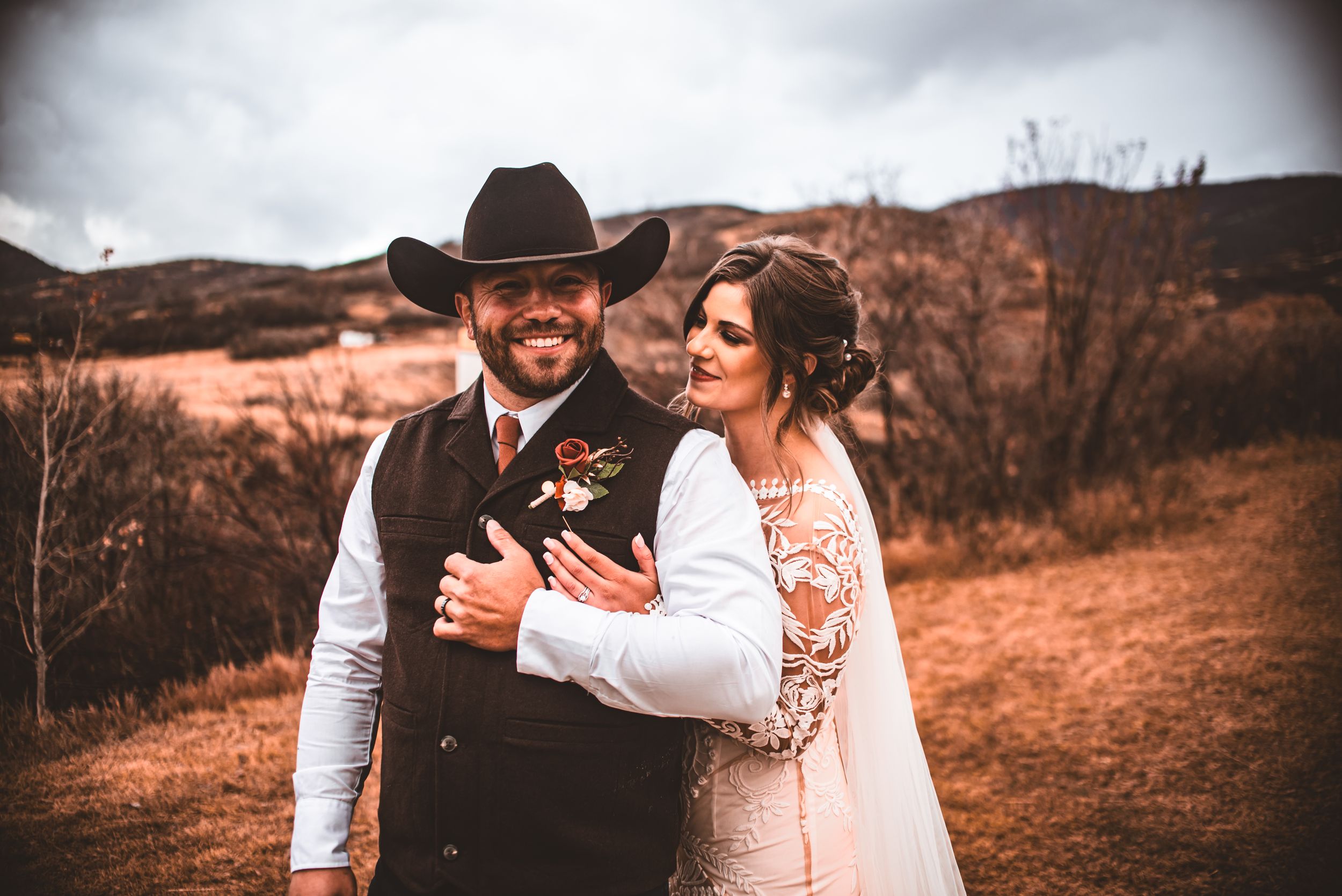 A couple in western wedding attire poses with their small dog against a desert landscape backdrop.