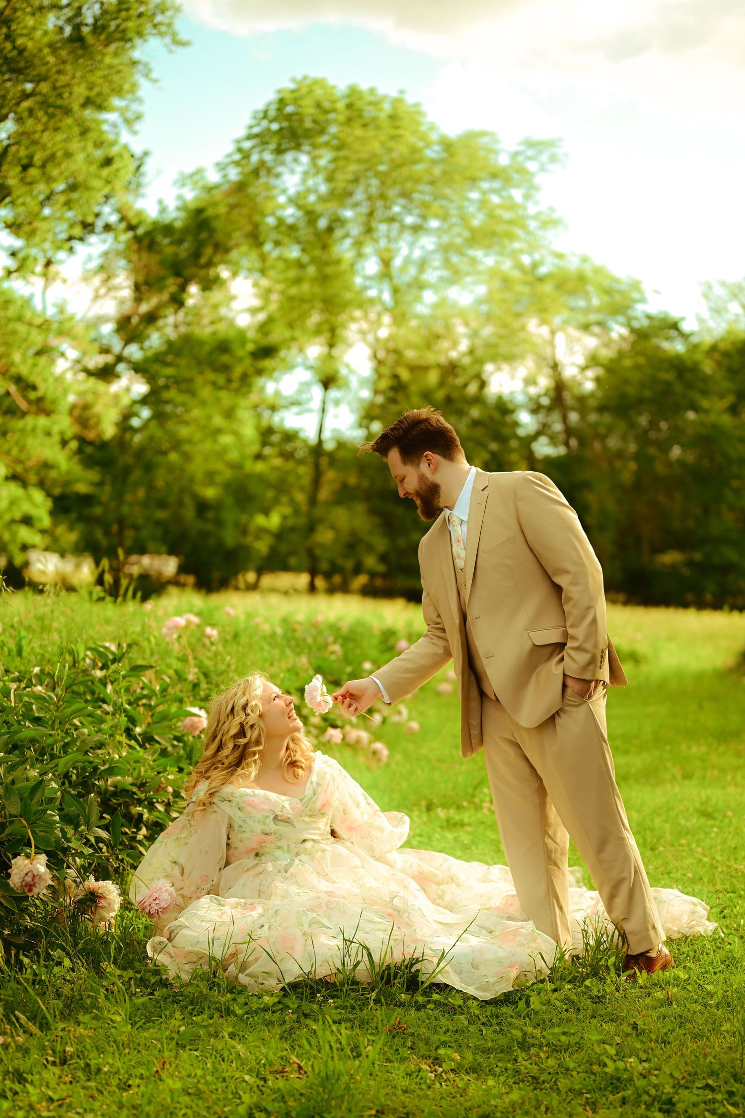 A wedding couple shares a romantic moment on lush green grass while her elegant white dress flows around them in the sunlight.