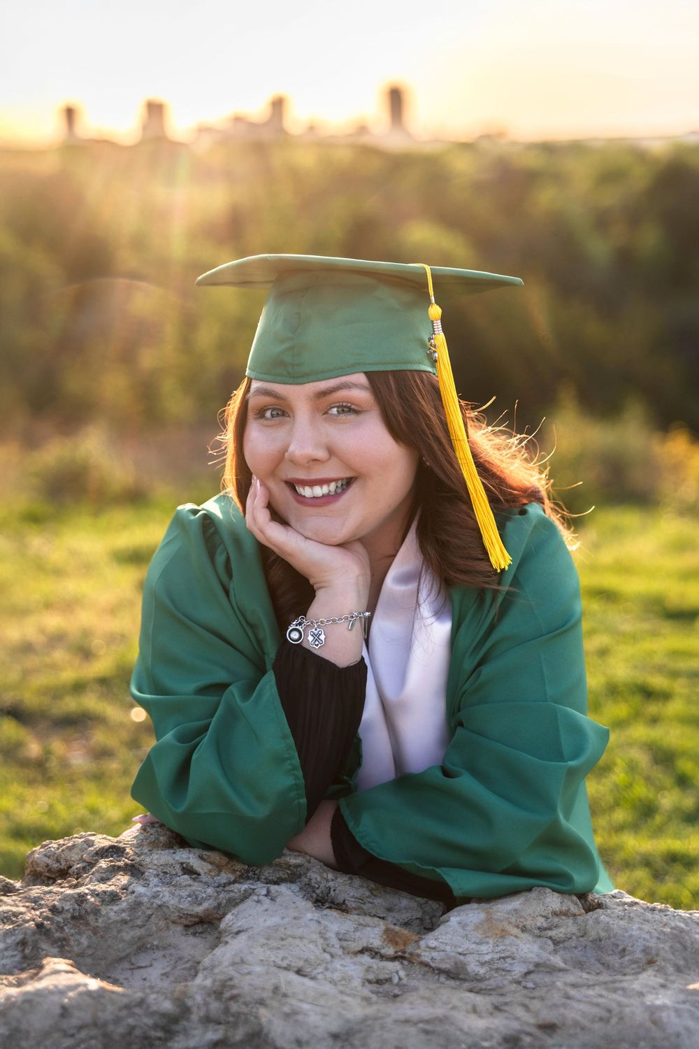A senior girl in her cap and gown poses on a rock with the Fort Worth skyline in the background.