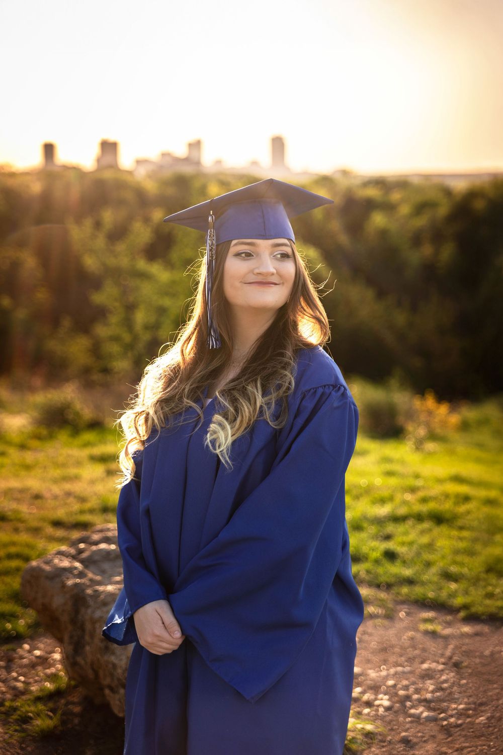 A senior in a cap and gown poses in the park with the Fort Worth skyline in the background.