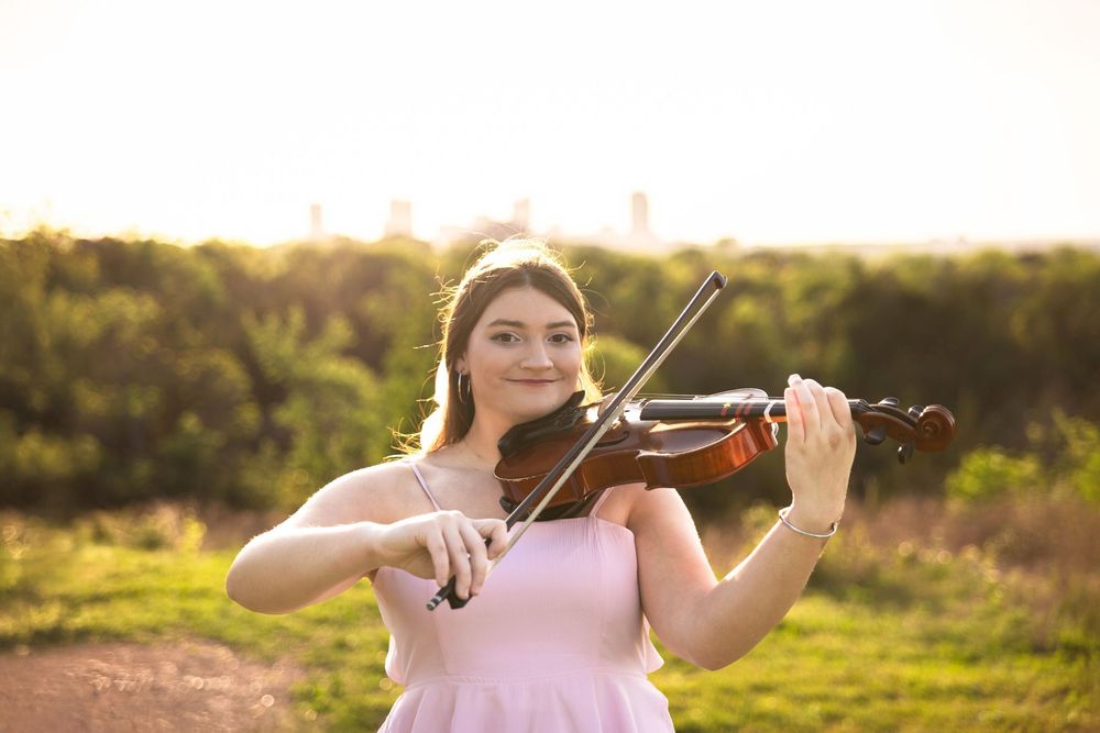 A young girl plays her violin in the park with the Fort Worth skyline in the background.