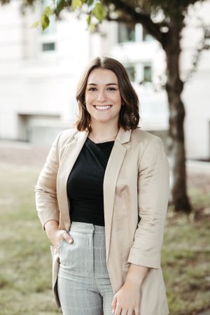Woman in beige blazer and black top smiles outdoors near building.